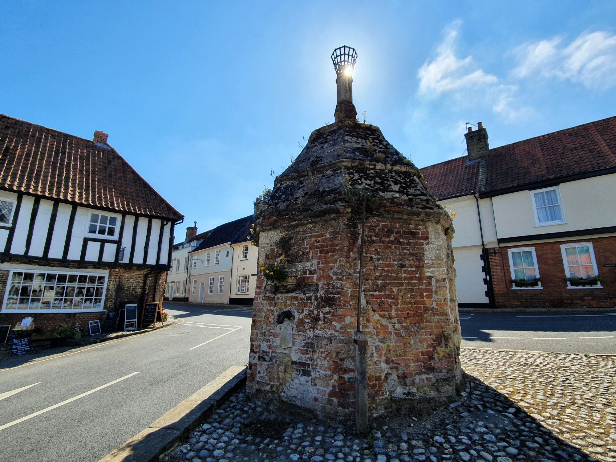 In foreground is the 16th century octagonal water pump made from brick in the middle of the village square. On top is a brazier, with the sun shining through it as if alight, casting a shadow in the foreground. In the background some old terraced cottages, the one on the left is timbered wattle & daub.