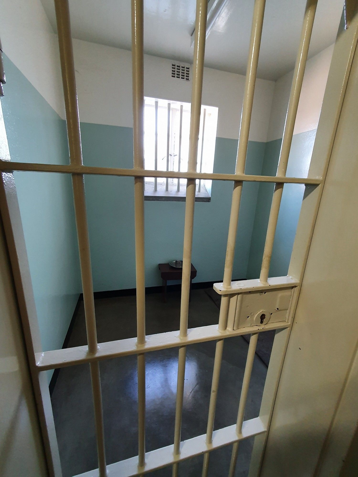 The photo is taken through the locked cell door made of vertical and horizontal metal bars through which the whole of the small cell is visible. The walls are painted pale green two thirds of the way up and then white. A small table against the far wall is the only furniture, with a small metal bowl on top. A large barred window above the table lights the cell as it is daytime, an old tube fluorescent light can just be seen on the ceiling.