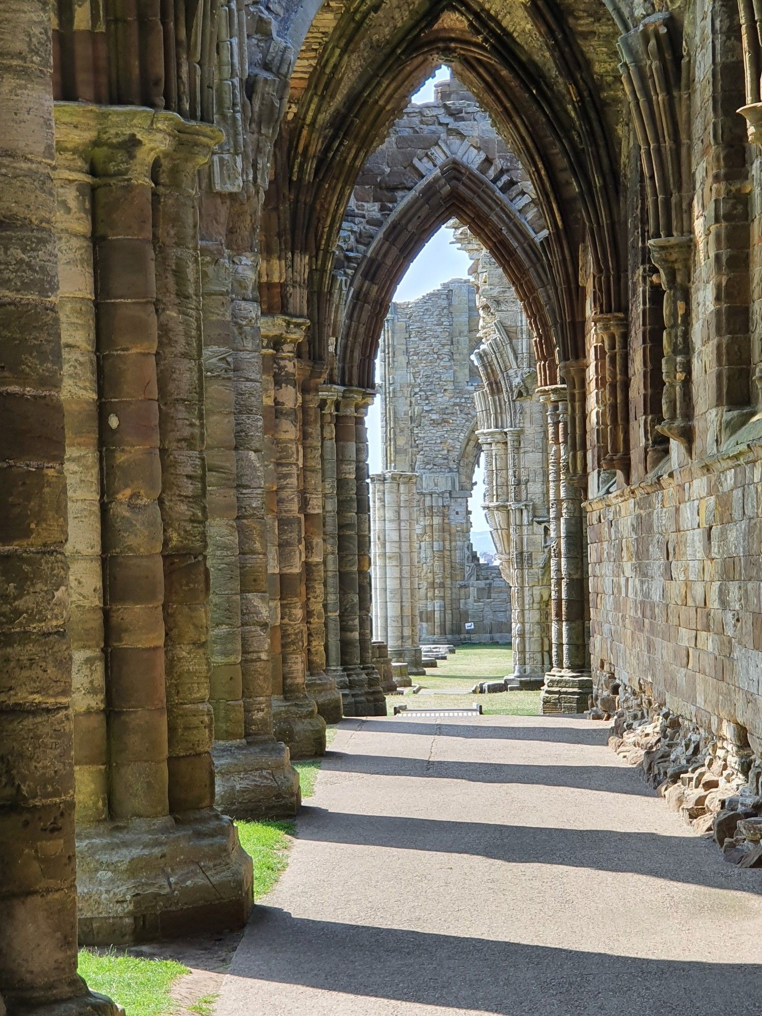 A view through a series of arches, part of the remains of Abbey on the headland at Whitby. The columns on the left cast a series of shadows across a path which leads to a ruined wall in the distance. A couple of houses in the town of Whitby can just about be seen in the far distance through a large window hole in the wall.