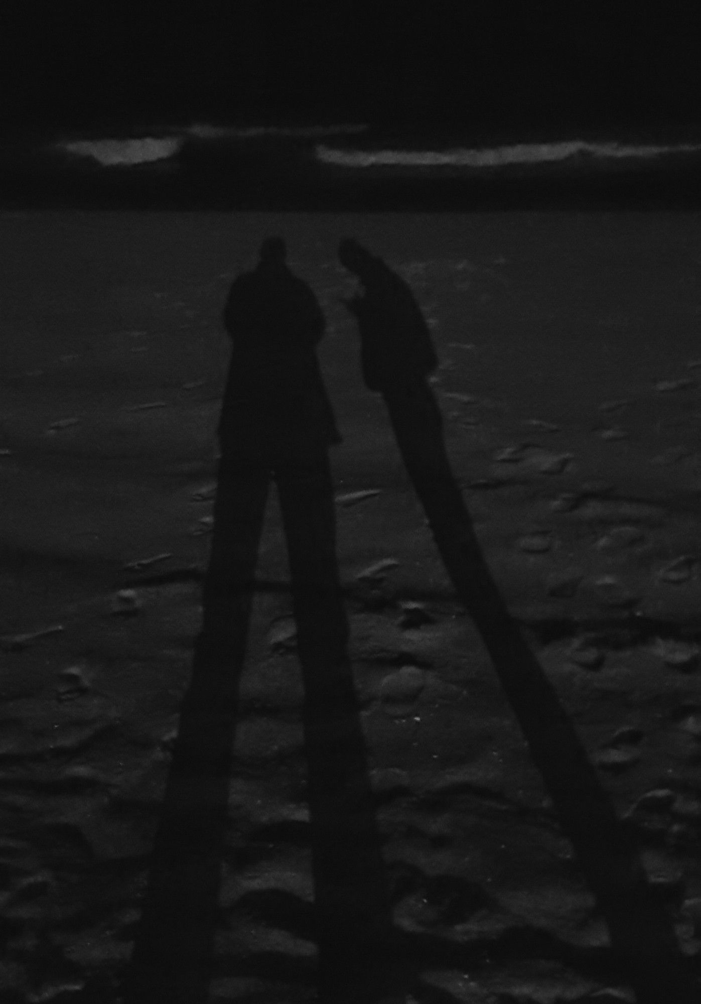 Photo at night-time on the beach. Shadow of two people lengthened across the sand, the shadow of one person taking the photo facing with 2 legs, the other square on with just one shadowed leg in showing. Footprints can just just be seen in the dark, wet sand. The white tops of some waves can be seen in the background.