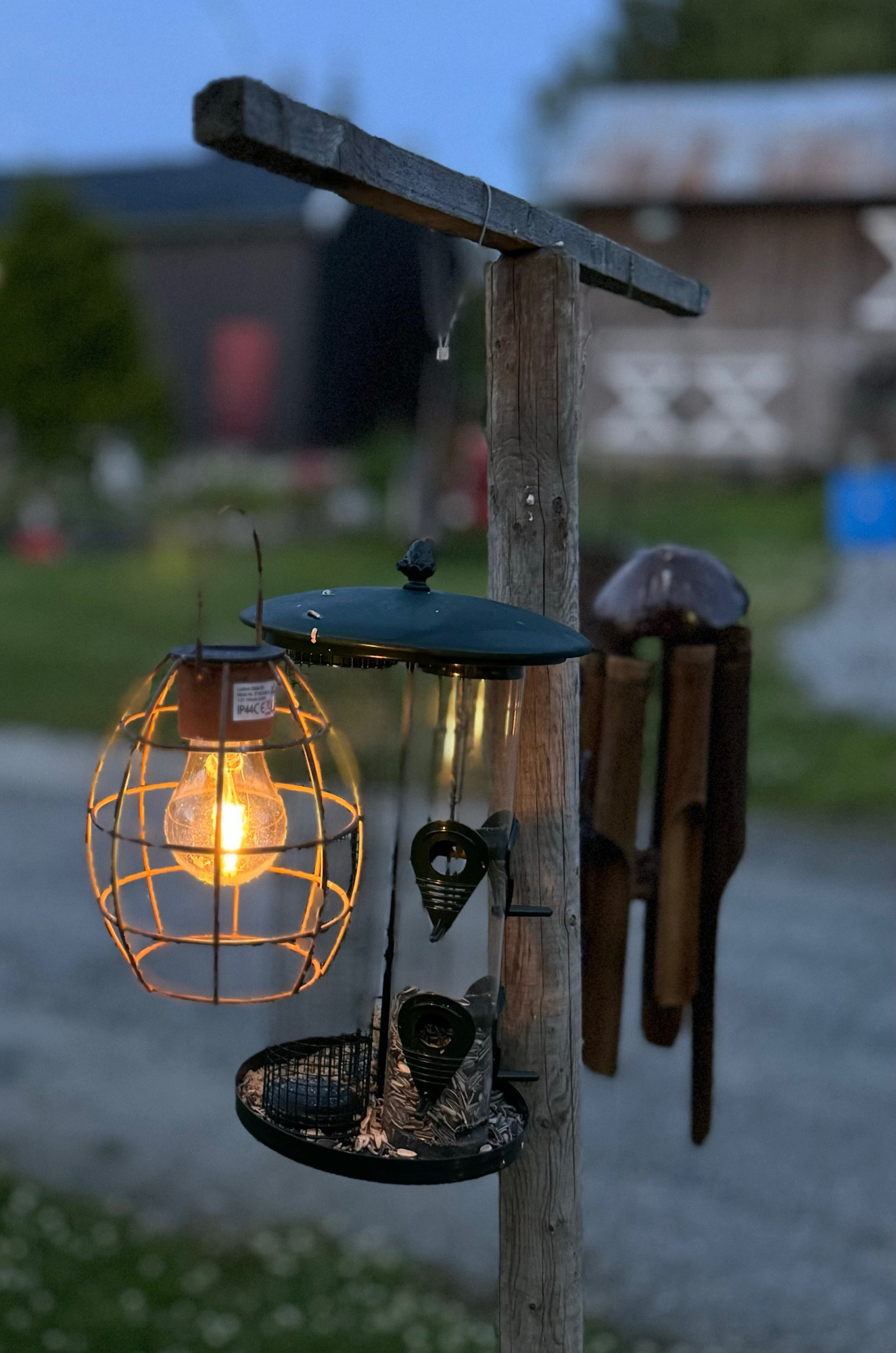 En liten solcelle lykt som lyser,en fuglmater og vindspill henger på fjøsmekka stolpe. Litt gras, låven og verkstedet uskarp i bakgrunnen. Tatt for litt siden i den lyse sommernatten