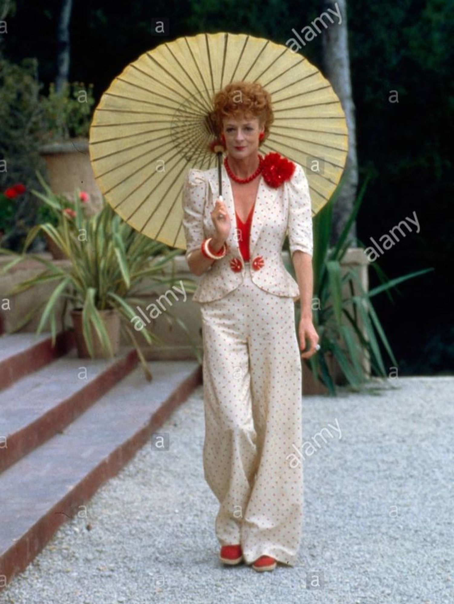 Maggie Smith wears a white with tiny red polka dot pants suit, pinched at the waist with two button, a cleavage showing red singlet under and a big red fabric at her left shoulder. No hat but she walks with a Japanese style parasol. a bangle and red espadrilles complete the look