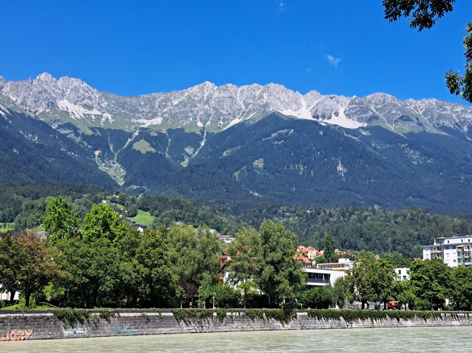 Das Foto zeigt eine Bergkette, darüber blauen Himmel, unten den Fluß Inn und dazwischen Bäume und Häuser.