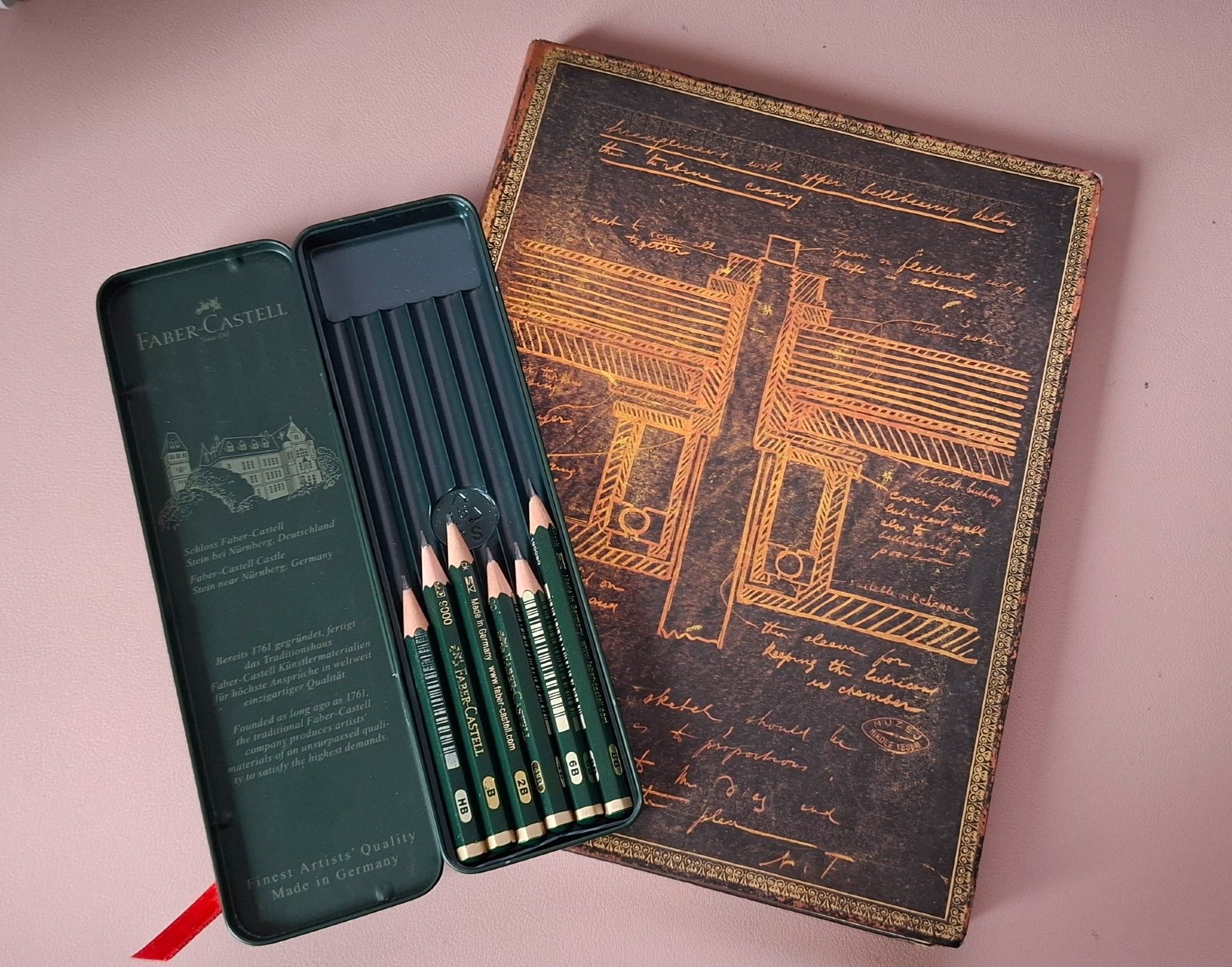 A photo of the sketchbook, with a pretty soft cover in browns and golds, depicting some of Tesla's inventions 
On top of it is the opened green case with the Faber-Castell graphite pencils from HB to 8B, most of them less than half their original size.