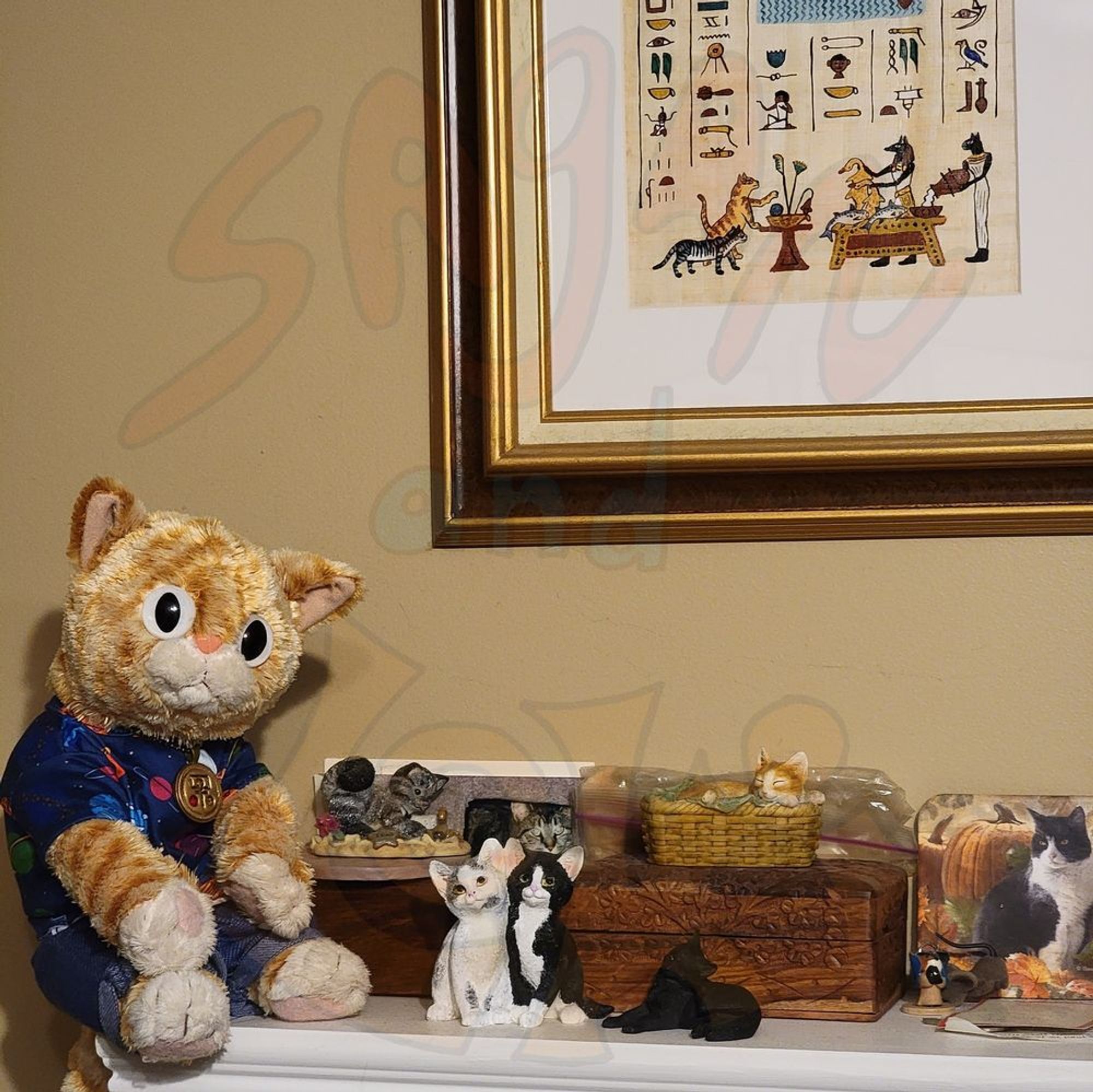 An orange plush cat sitting on a mantel next to figurines of cats and under an image of Bast and Anubis offering food to cats in the afterlife.