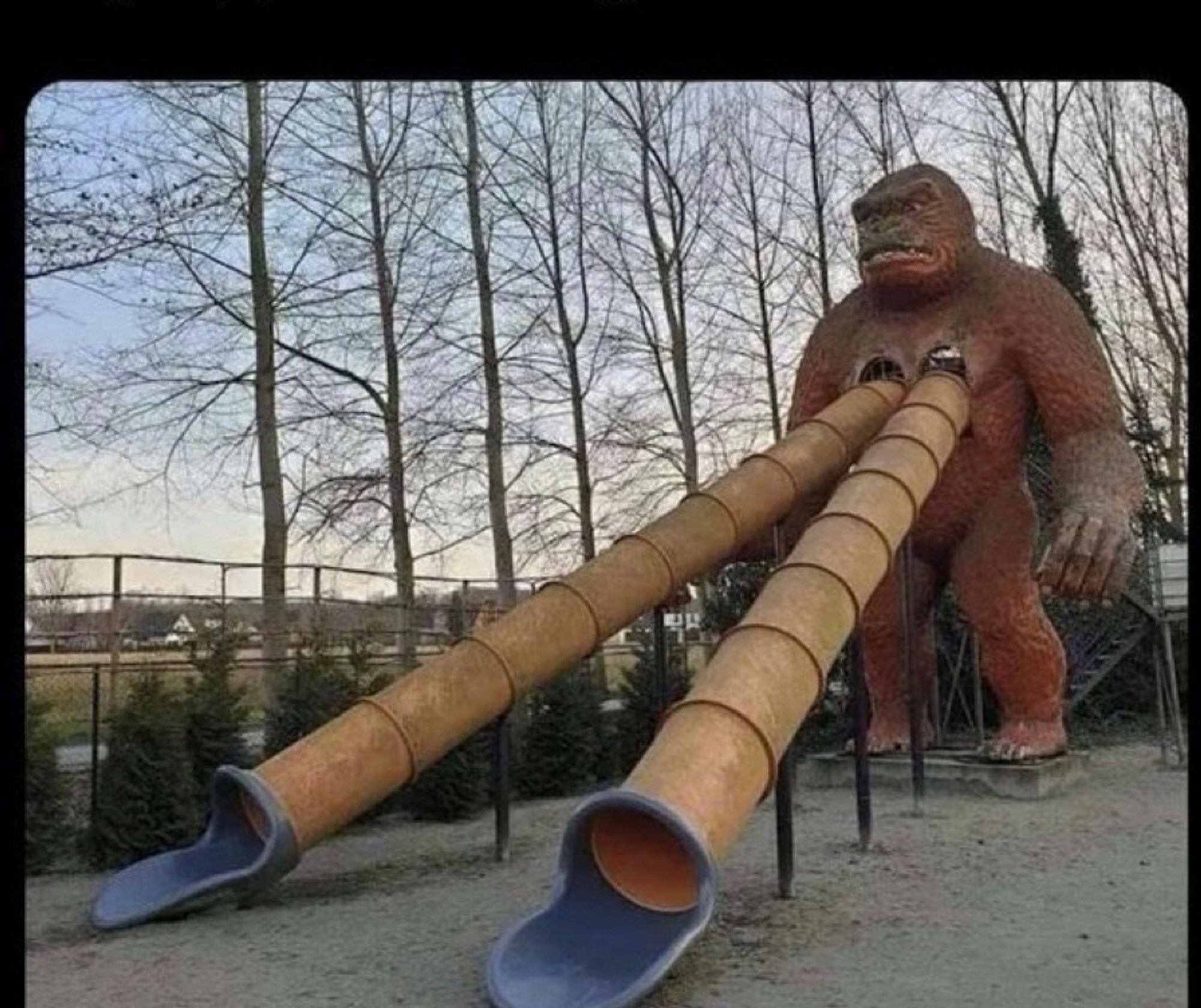 A park attraction where a three story tall brown gorilla stands with two straight slides coming from where both its nipples are. The slides are dirty orange with blue ends.