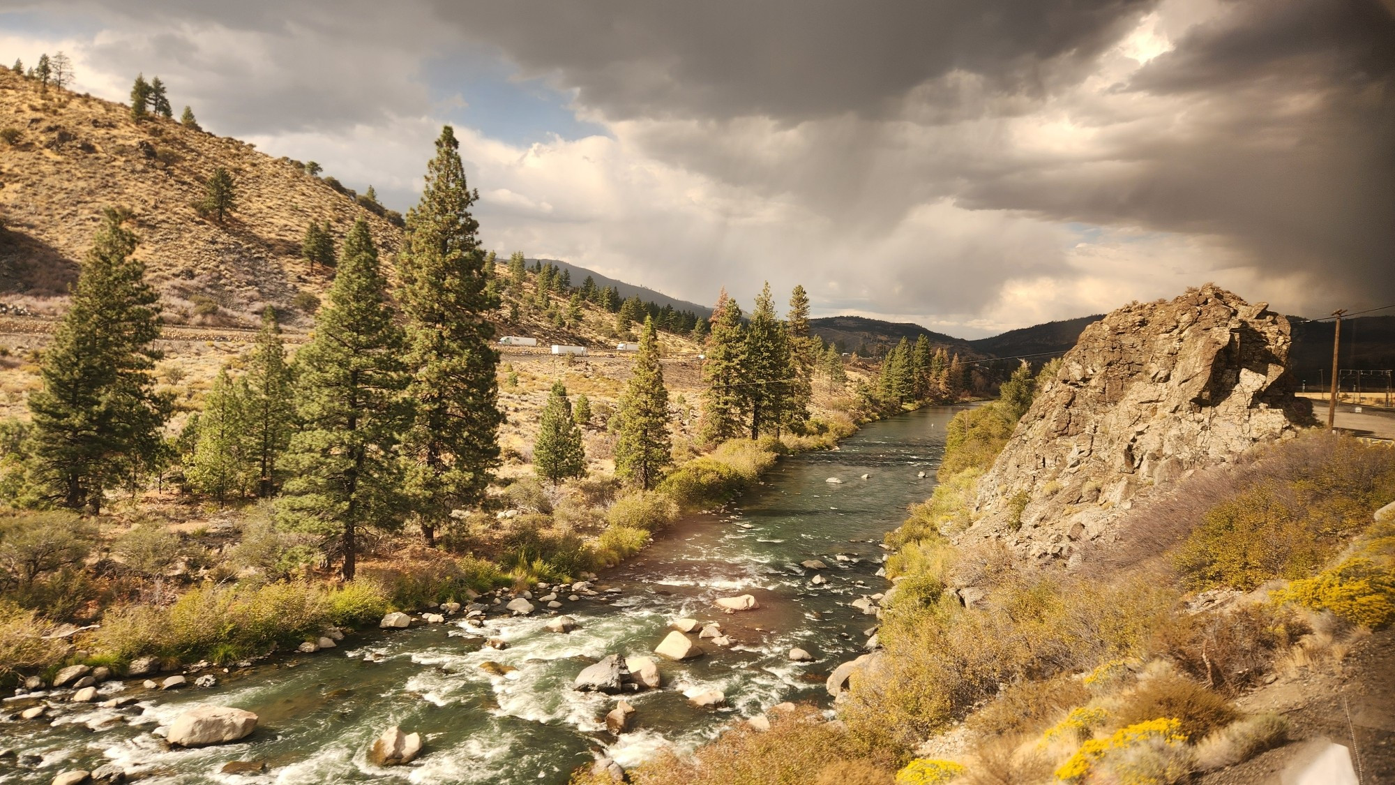 A small river is running through a mountain landscape. There are rapids in the foreground and evergreen trees on the left side. The sky has mostly dark clouds with one small patch of blue.