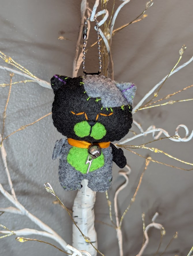 Franken-Neko has black head with another Neko's gray head sewn on. It also has green cheeks, a green tummy, one black arm and one gray arm. It has a bell and lightning bolt tied around its neck with an orange ribbon. Stands roughly 3” x 1.5” / 7 x 4 cm from ears to feets.