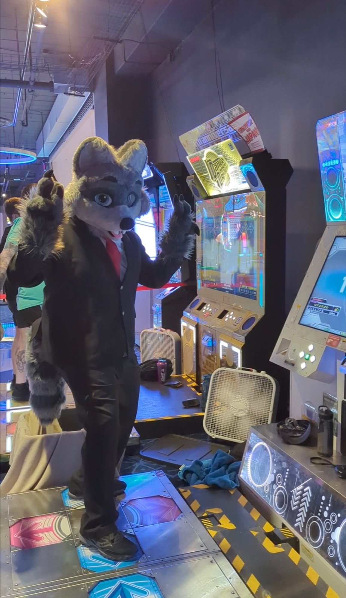 A photo of someone in a partial raccoon fursuit  with a suit jacket and suit pants on. He's standing on a Dance Dance Revolution pad facing the camera and giving two peace signs.