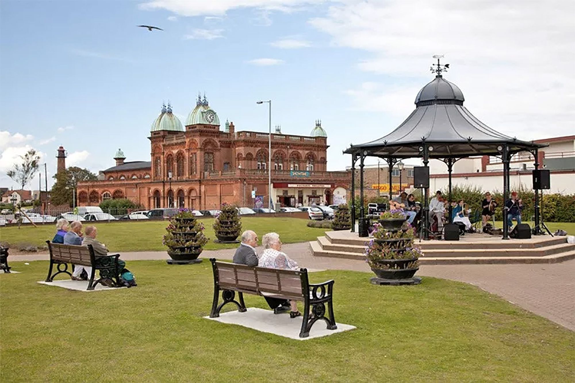 An image of the Gorleston Pavilion Theatre and Bandstand. There are people sitting on benches listening to some musicians playing on the bandstand. The Theatre can be seen in the background.