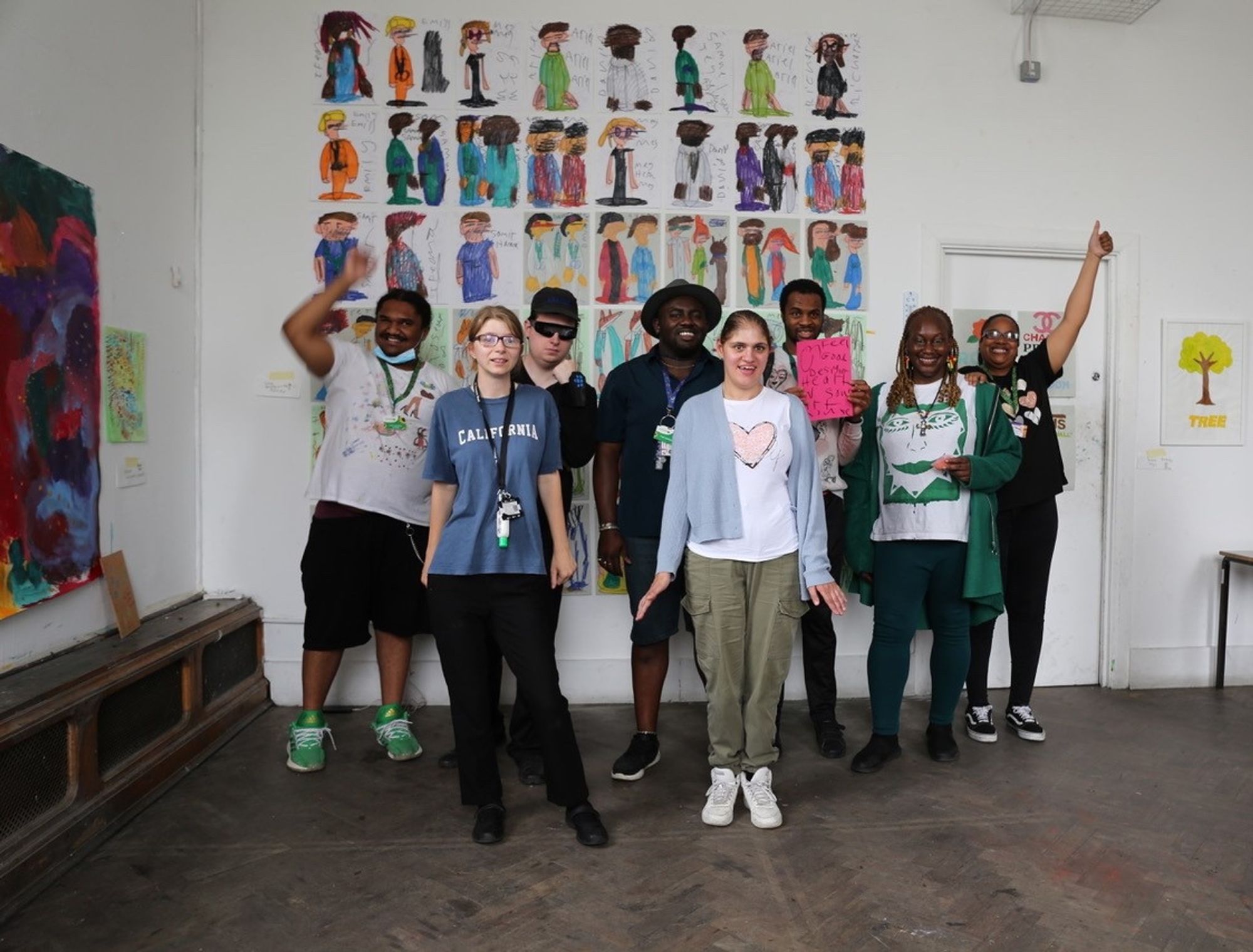 A workshop run by Heart n Soul, an organisation that works in partnership with Lewisham Arthouse at the Old Deptford Library. Eight people are seen standing in a line, smiling for the camera. Behind them is a wall of paintings.  Credit - Richard Phoenix.