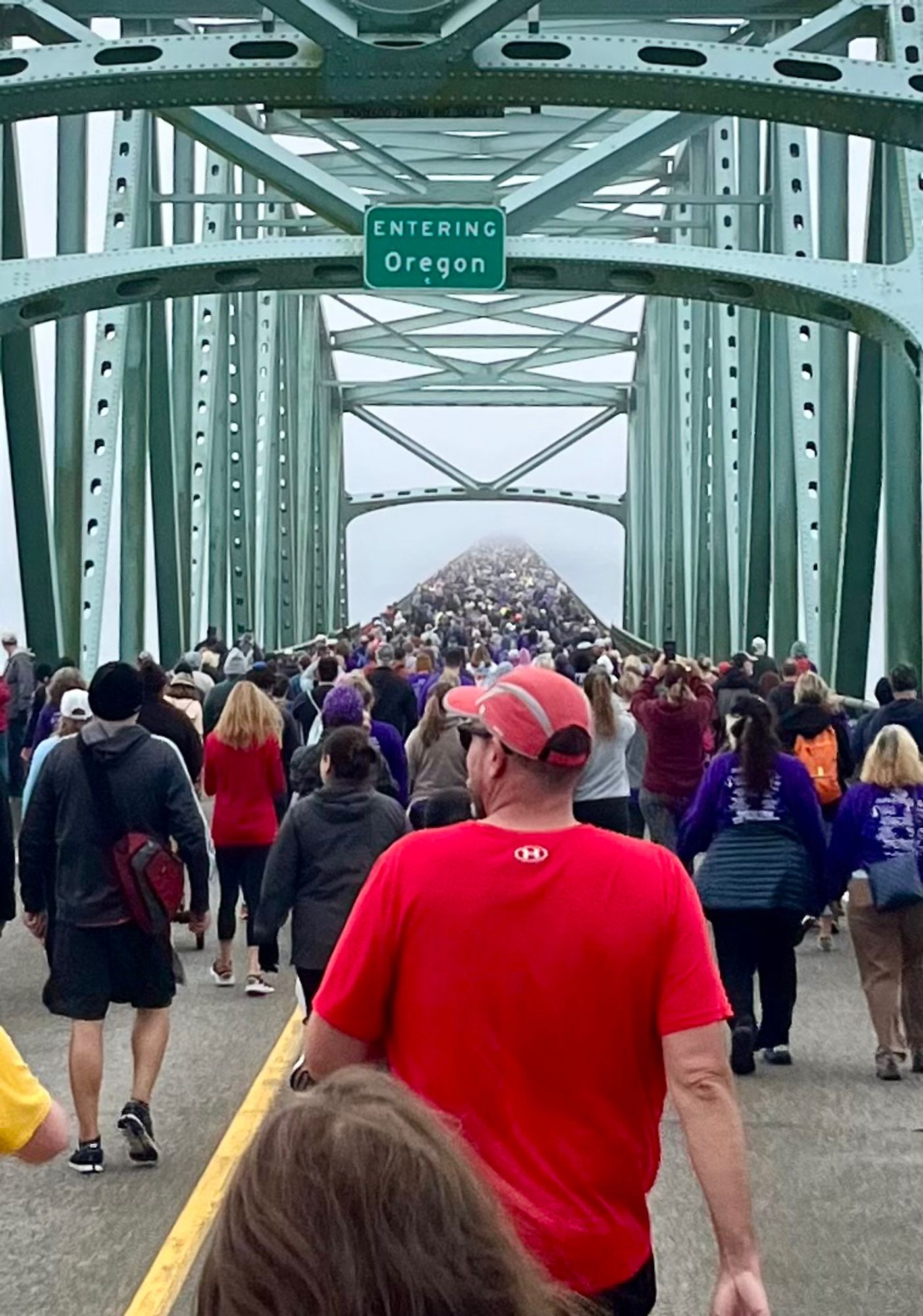 Hundreds of walkers on a bridge pass under a sign on the bridges iron superstructure that says “Oregon.” It’s foggy in the distance, it looks like they are walking into nothingness.