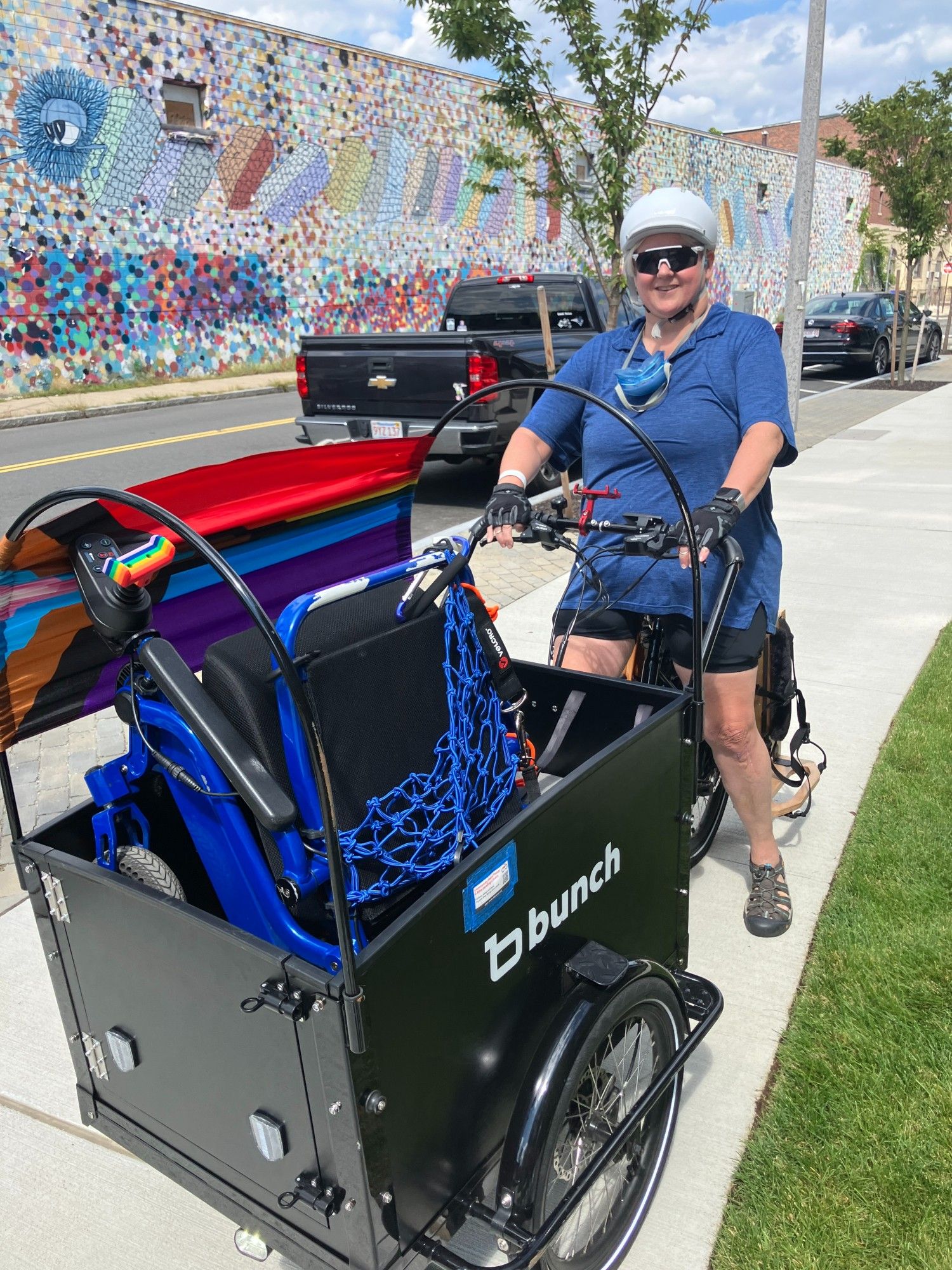 A person wearing a bicycle helmet and sitting on the seat of a cargo trike. The front-loading cargo trike is labeled "Bunch" on the side, and contains a folded up wheelchair.