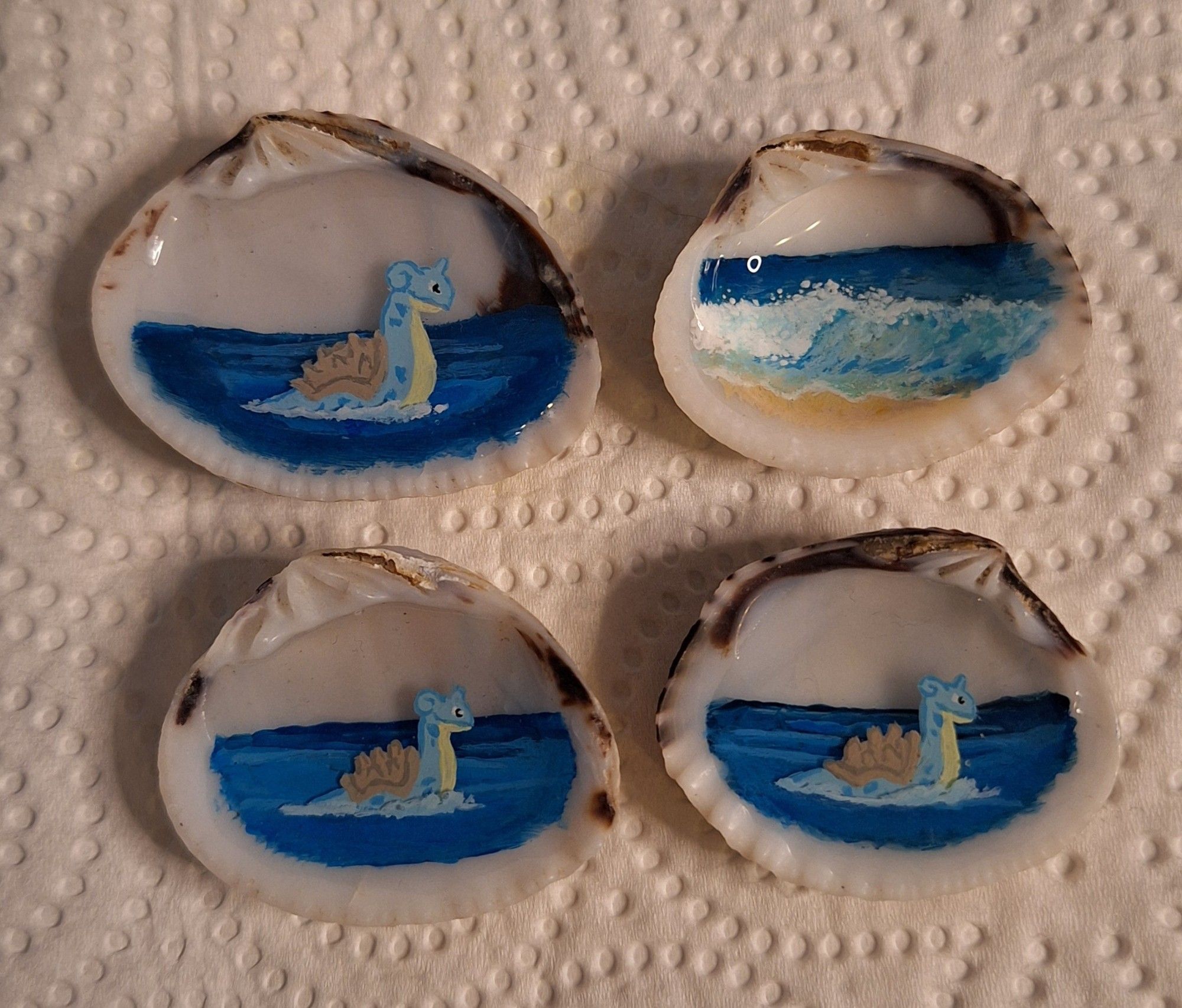 4 small painted shells, of which 3 are painted with a Lapras Pokemon swimming in the ocean and 1 is painted with an ocean wave