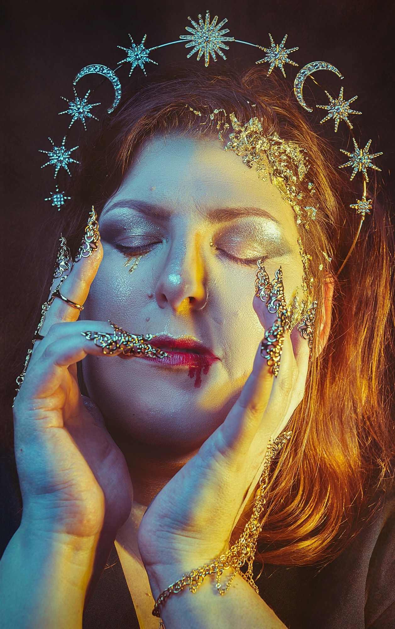 Close up studio portrait where the model has her eyes closed with gold and silver makeup accents catching the light, is wearing a sun and moon tiara and has her hands beside her face with golden jewelry