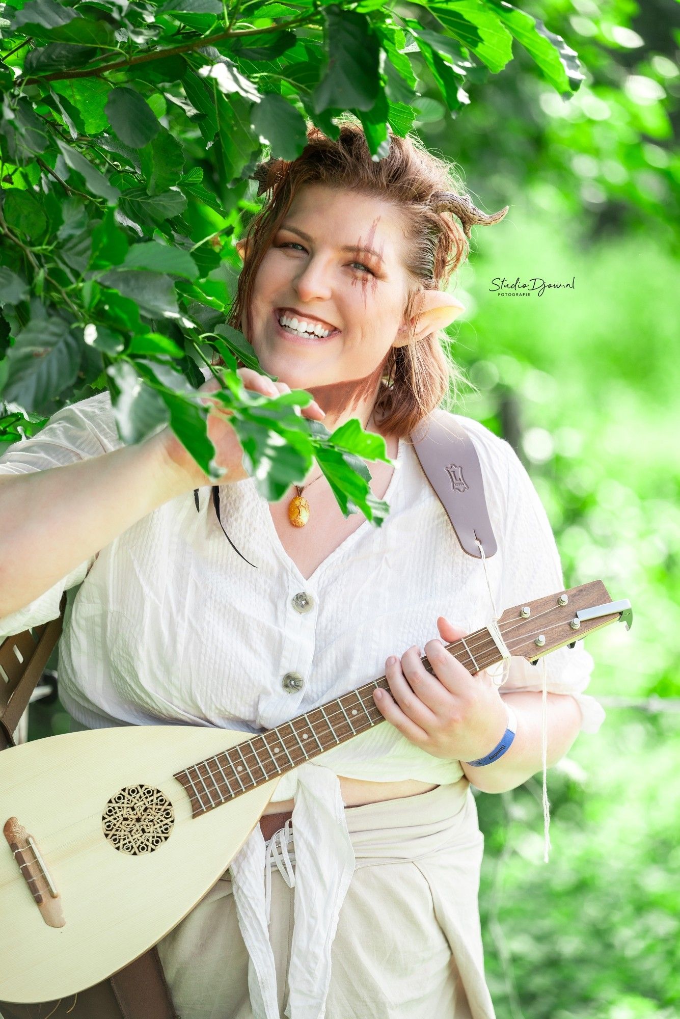 A faun oc cosplayer smiling, pushing away leaves from a tree while holding a ukelele that looks like a lute