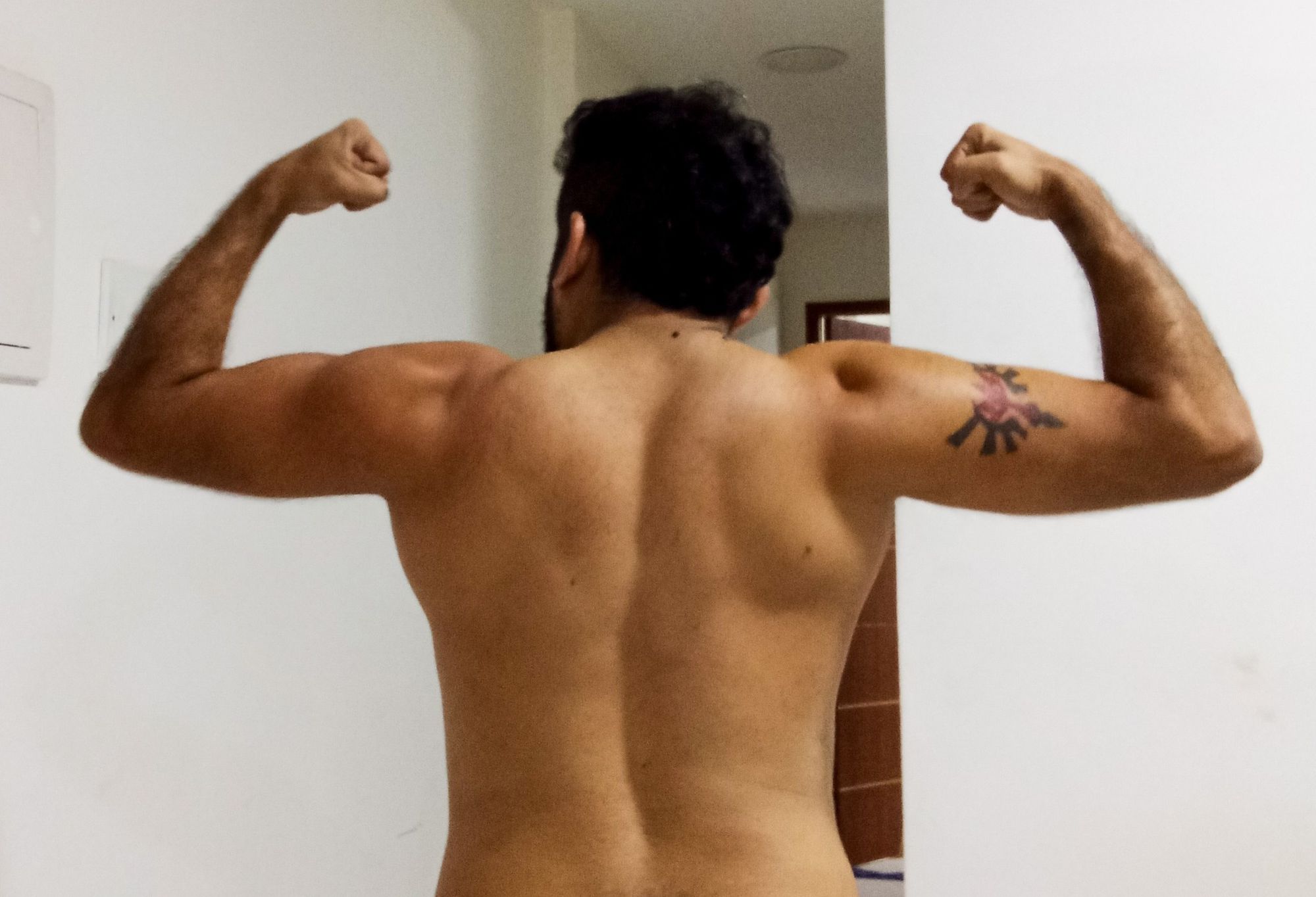 A foto mostra um homem pardo sem camisa, de costas, flexionando os braços para mostrar os musculos das costas e dos braços.
No braço direito, abaixo do ombro, o homem tem uma tatuagem de Zelda que mistura o simbolo de Hyrule com o símbolo dos Sheikah.