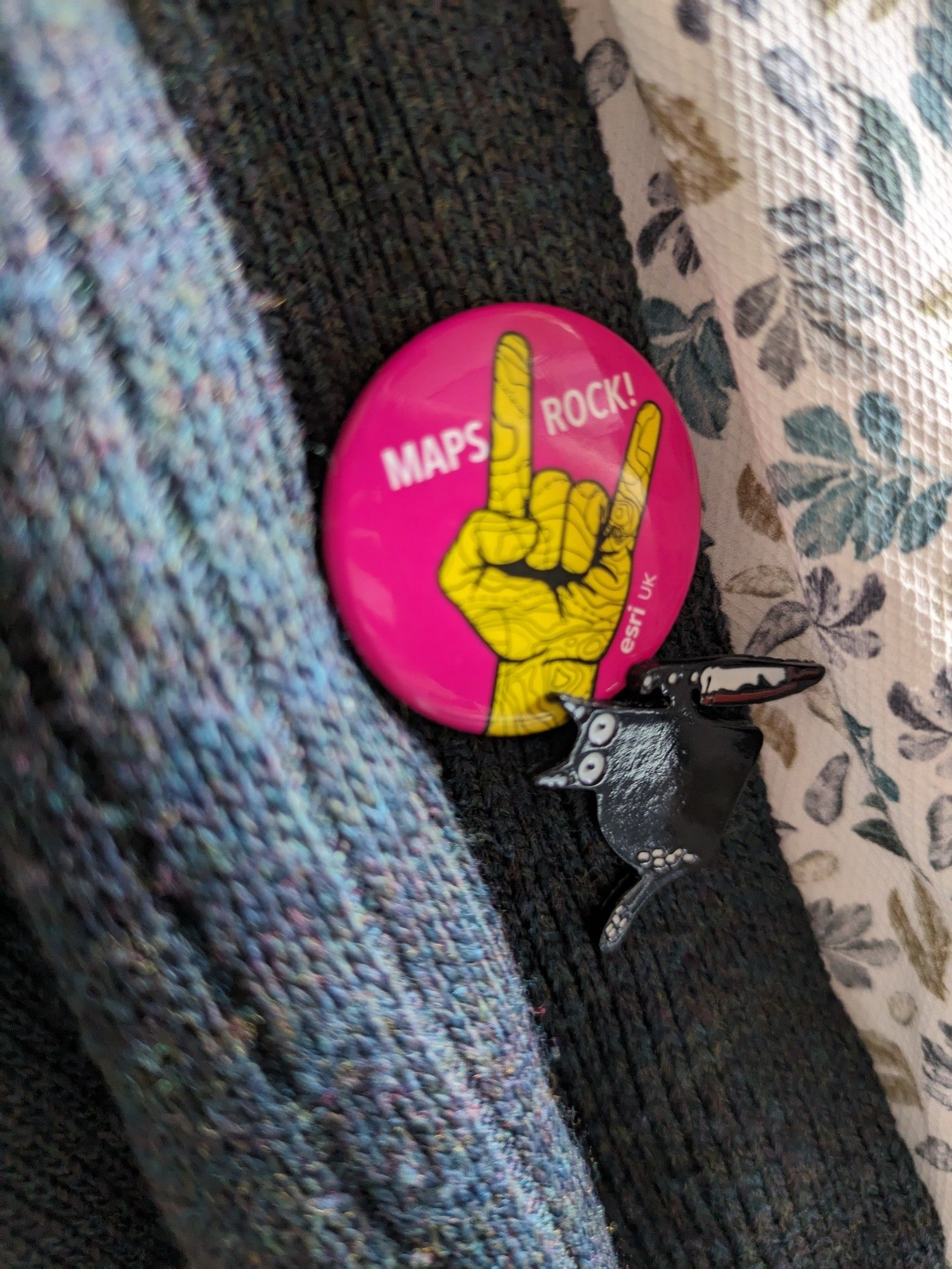 A pink pinbadge showing a hand in the horns position stylised to look like cartographic markup