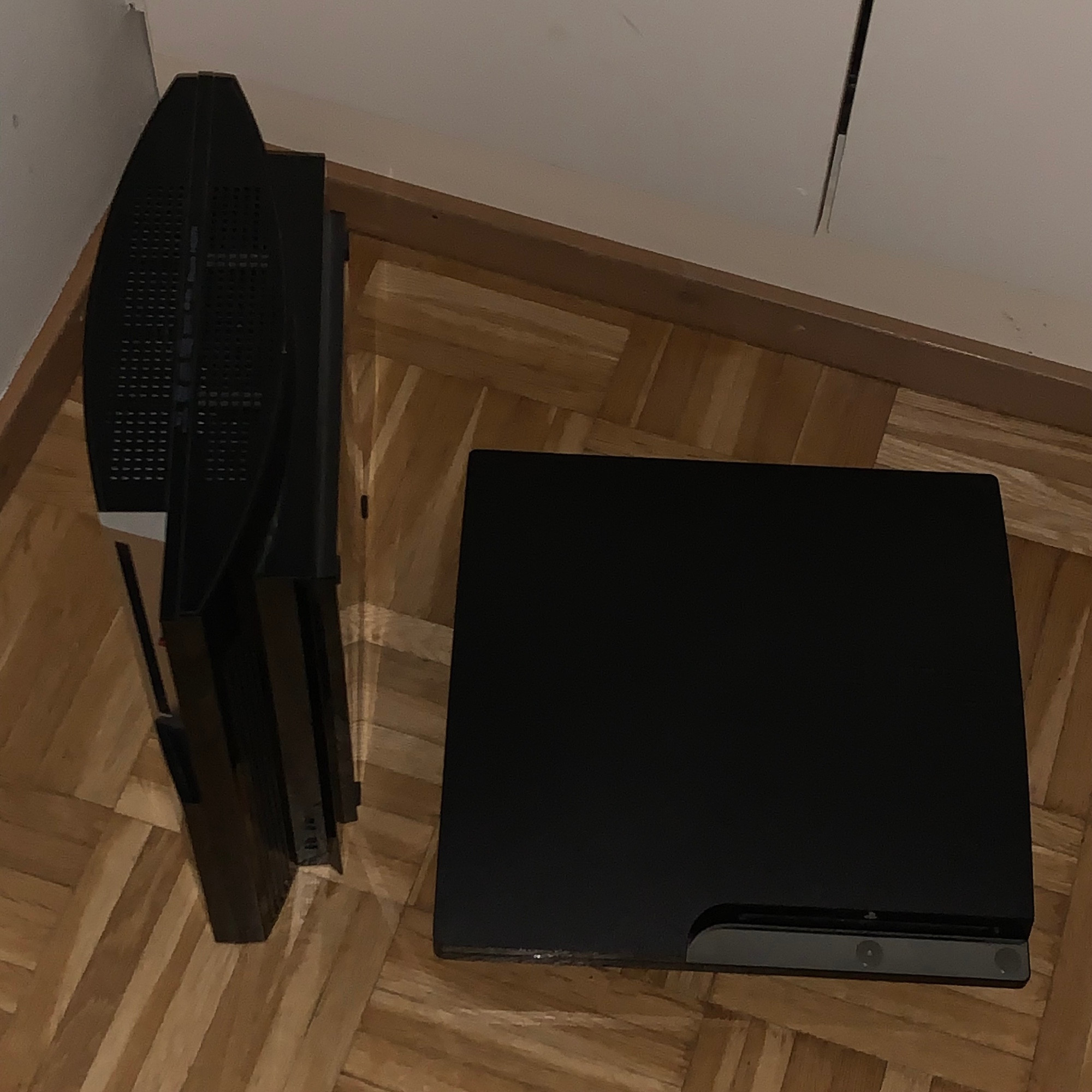 a fat model PS3 stood up vertically and a slim model PS3 laying horizontally in a dim room lit by camera flash, top down view, the slim model’s lettering on the top cover saying ”PS3” has been lost