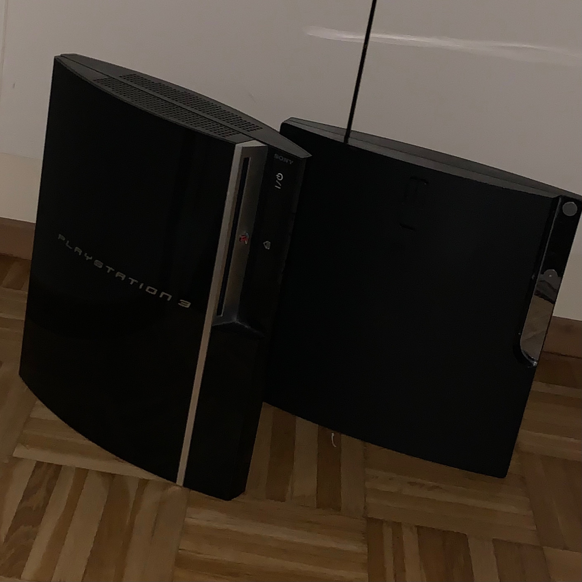 a fat model PS3 and a slim model PS3 stood up vertically in a dim room lit by camera flash, fat model PS3 has shiny lettering saying ”PLAYSTATION 3”