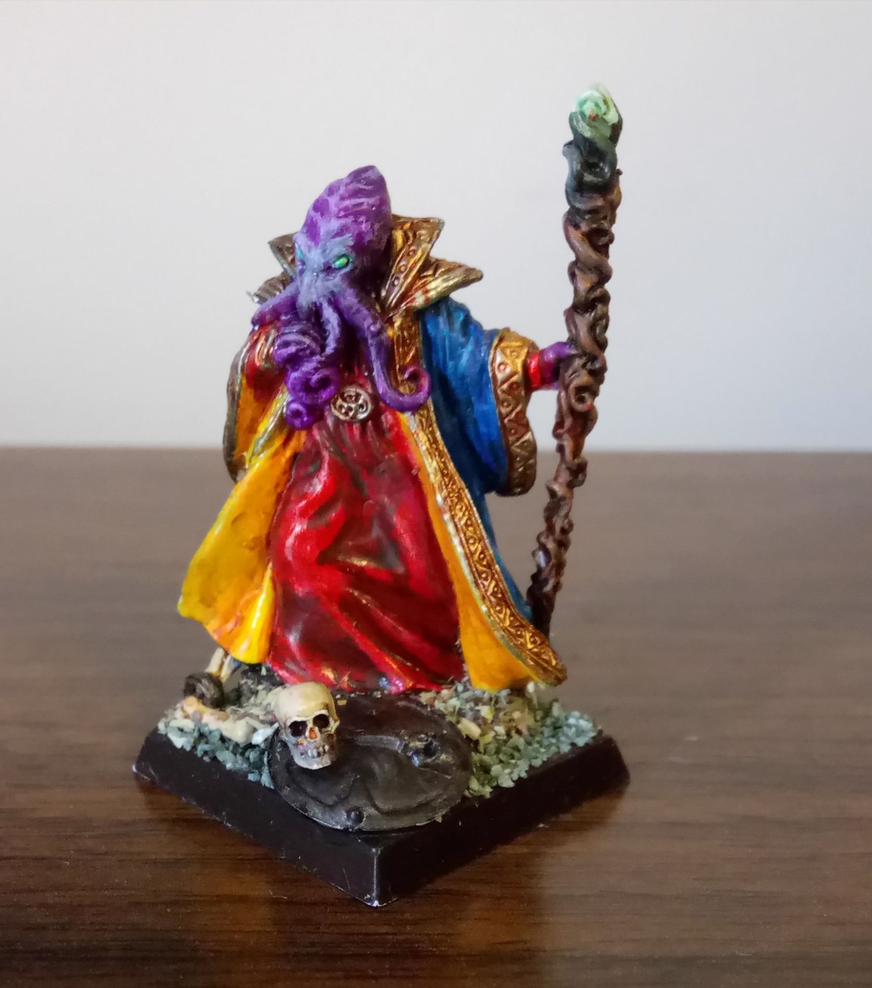 Purple-skinned Mindflayer with glowing green eyes in royal blue robe with gold trim and yellow lining.  He is holding a twisted wooden staff with a glowing green stone at the top.  There is a skull and shield by his feet.