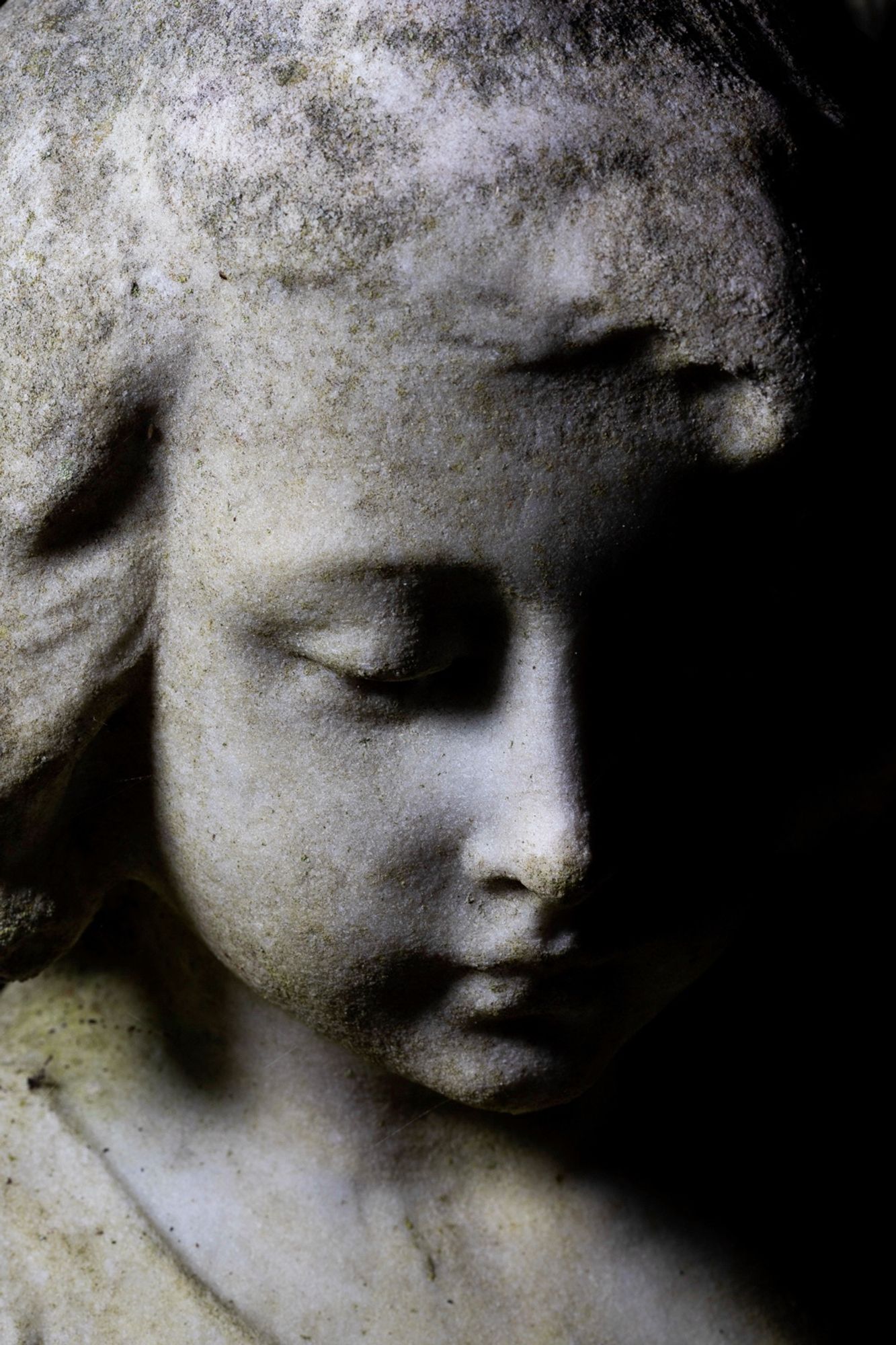 Photograph of a statue in a cemetery. Close up of the face. The face is of a child, a girl, an angel. She is looking down in reverence or mourning. There is dramatic lighting so half the face is in shadow. “like a Joy Division album.”