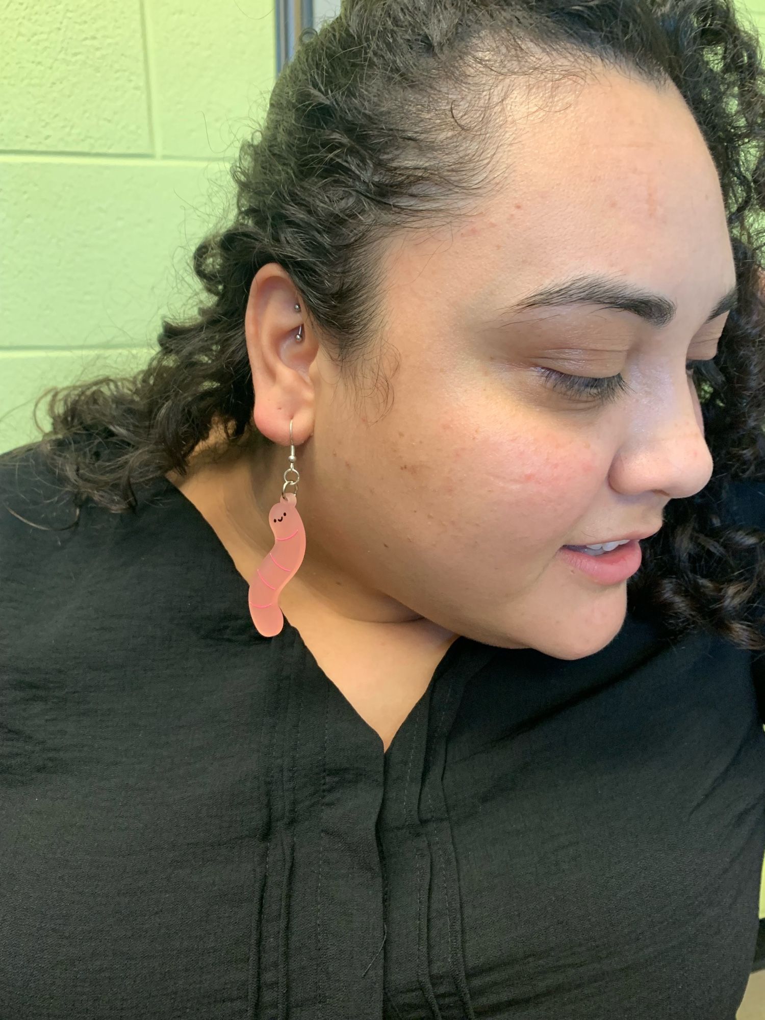 Andrea is wearing a black shirt and is holding her long, black curly hair to show off her earrings. They are large, pink worms