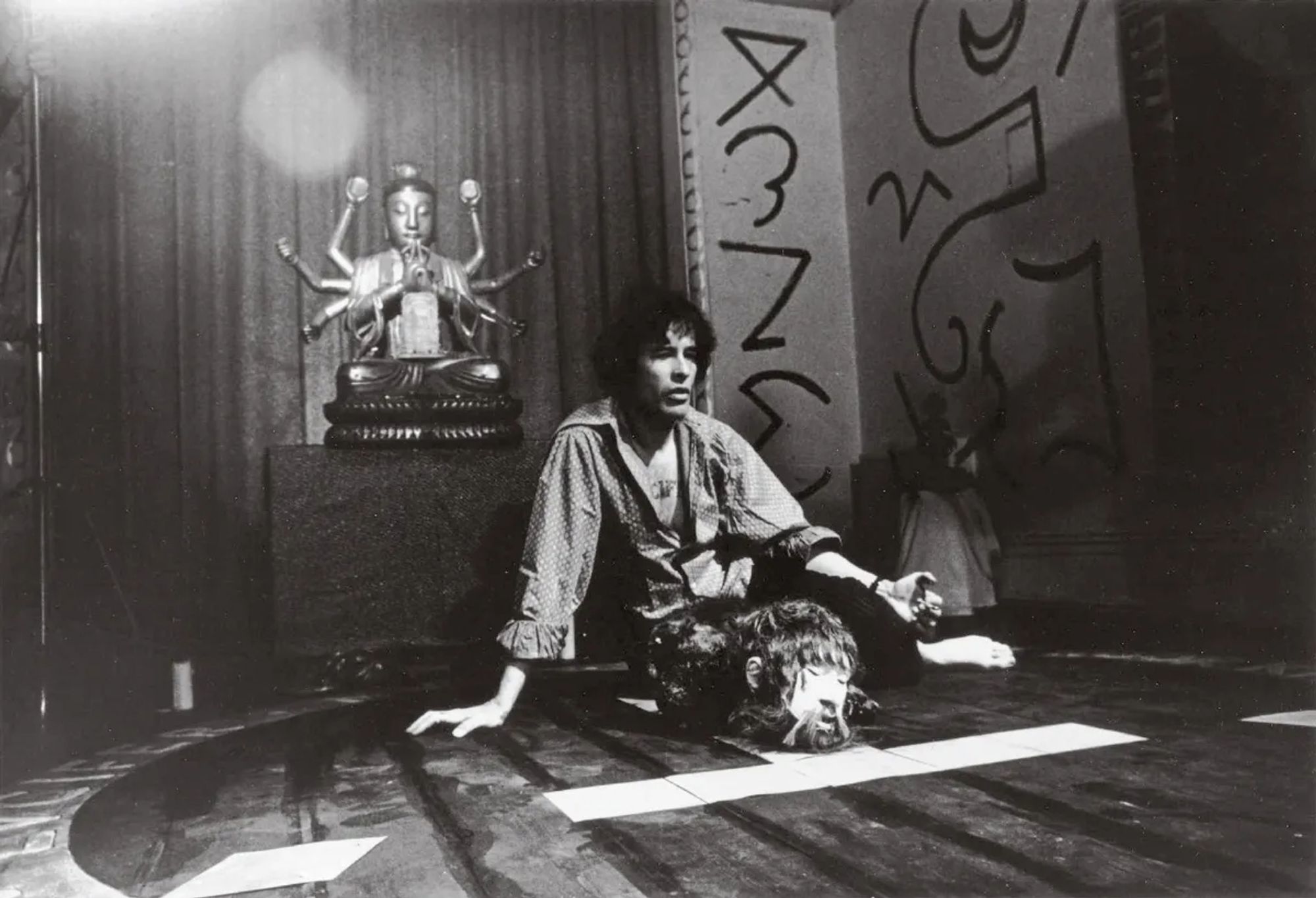 A young Kenneth on the set of Lucifer Rising.  In the background is a multi-armed Buddha statue and a wall decorated with a variety of sigils.  Anger sits at the center of a circle, arranged in the general outline of the A.'.A.'. temple, with a fake severed head on the Tau.