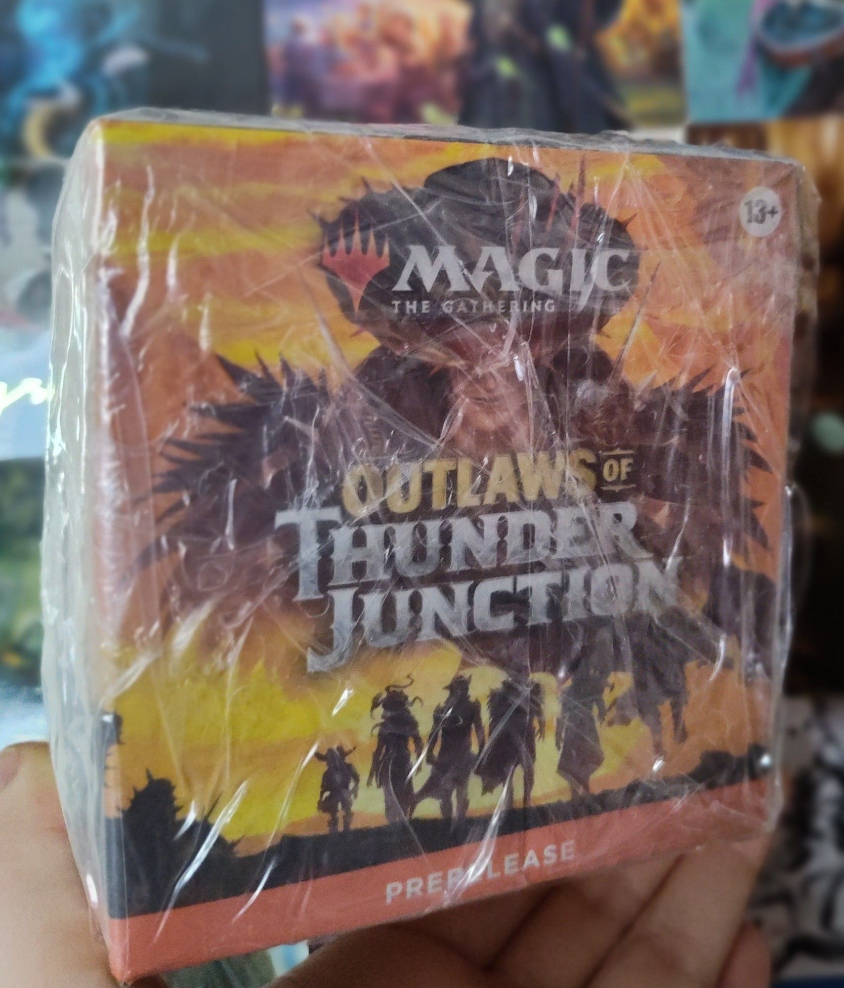 A vibrant yellow / orange sunset box with the words "Magic the Gathering" at the top along with their logo. Behind the words is a person's face. They have spikey hair and they use shoulder pads that also have spikes. In the middle it says "Outlaws of Thunder Junction" with a group of 5 people walking towards the sunset. Underneath that it says "Prerelease".