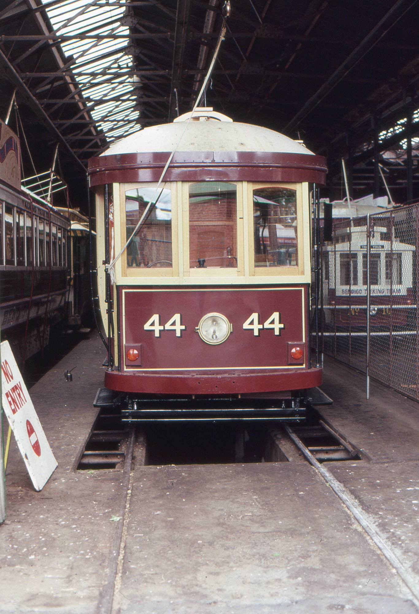 A front-on view of a WW1-era bogie tram in maroon and cream livery.  The drivers cab has three windows, and a single large centrally mounted headlight is on the apron.  The tram is in a tram shed with other trams visible either side  of it, including, to its right, a half or one third scale mini tram that was used to promote Bendigo Tramways.