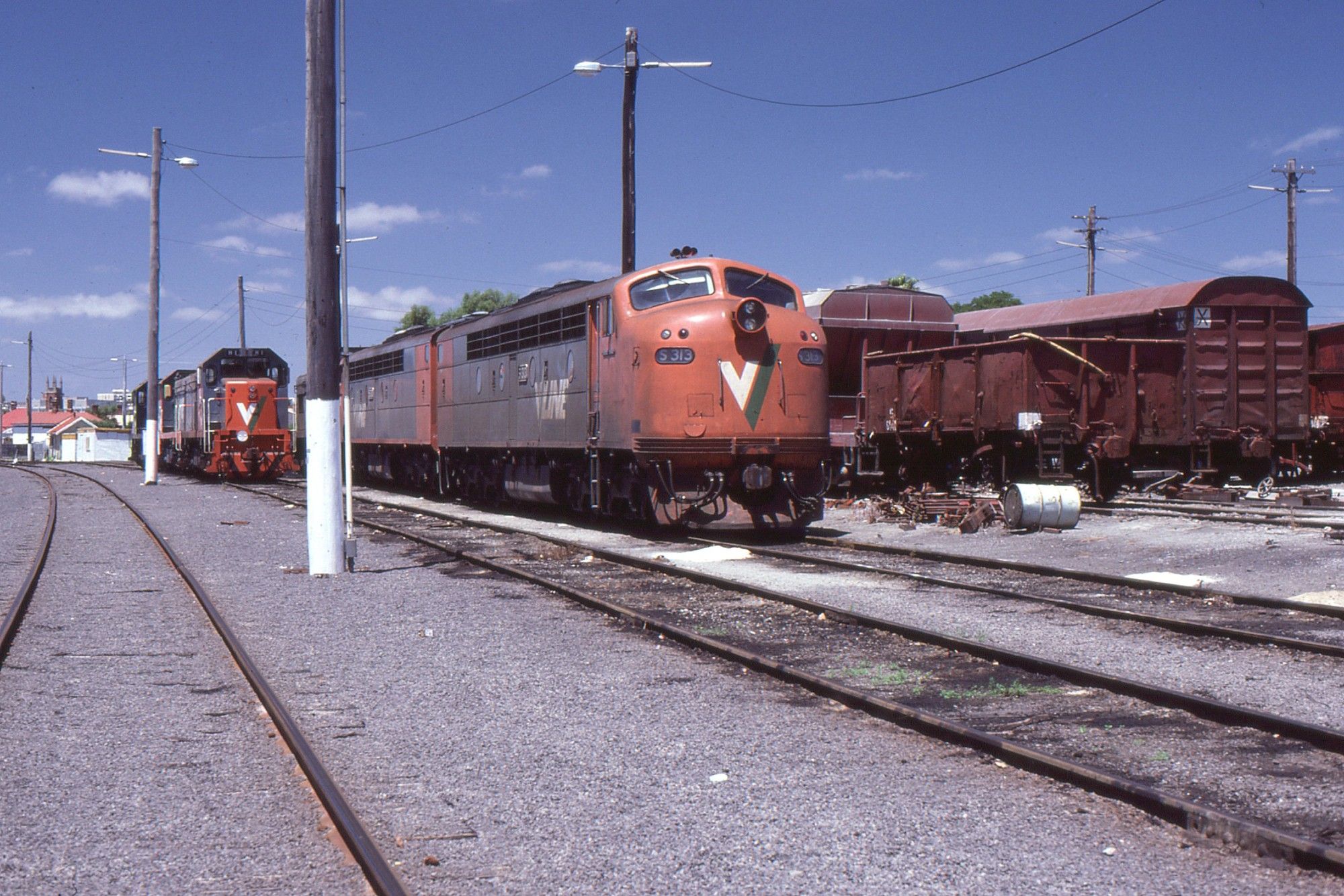 On a bright sunny day beneath a blue sky we see a streamlined EMD engined cab unit diesel locomotive in orange and grey VLine livery with white and green V/ logo on its nose, sitting on a track in a locomotive depot.  Attached behind it is a similar loco, and on the adjacent track to the left in the background are three hood unit diesel locos.  On more tracks to the right is a collection of dark red liveried goods rollingstock including an open wagon a box van and a hopper wagon.