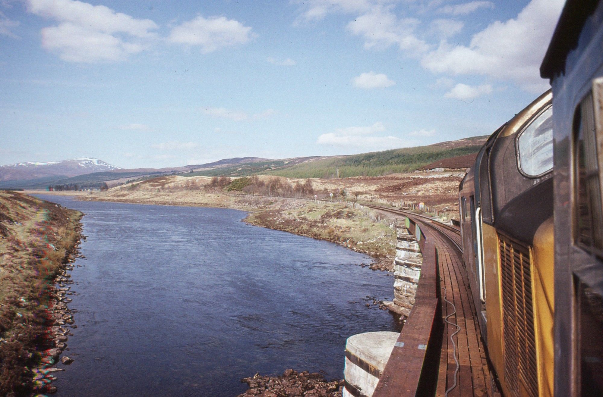 A view taken looking forward through an open window on the left hand side of a passenger train.  We are in the front carriage immediately behind the diesel locomotive, the rear cab of which is partly visible at right just a couple of metres in front of us.  The train is crossing a curved bridge over a waterway which is filled with dark water.  The countryside either side of the waterway is a barren highland landscape.  In the distance on the horizon is a snow capped peak beneath a blue sky flecked with scattered white clouds.