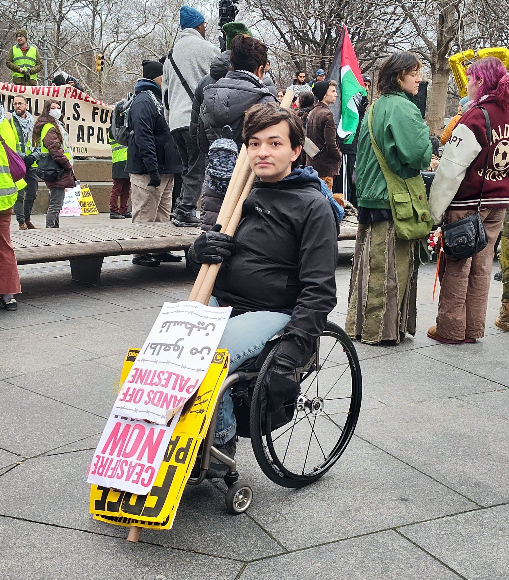 an activist in a manual wheelchair carrying a good quantity of protest signs and they are holding them upside down as they make their way around the rally to see who needs to signs. The signs have pro Palestine messages on them like Ceasefire now!