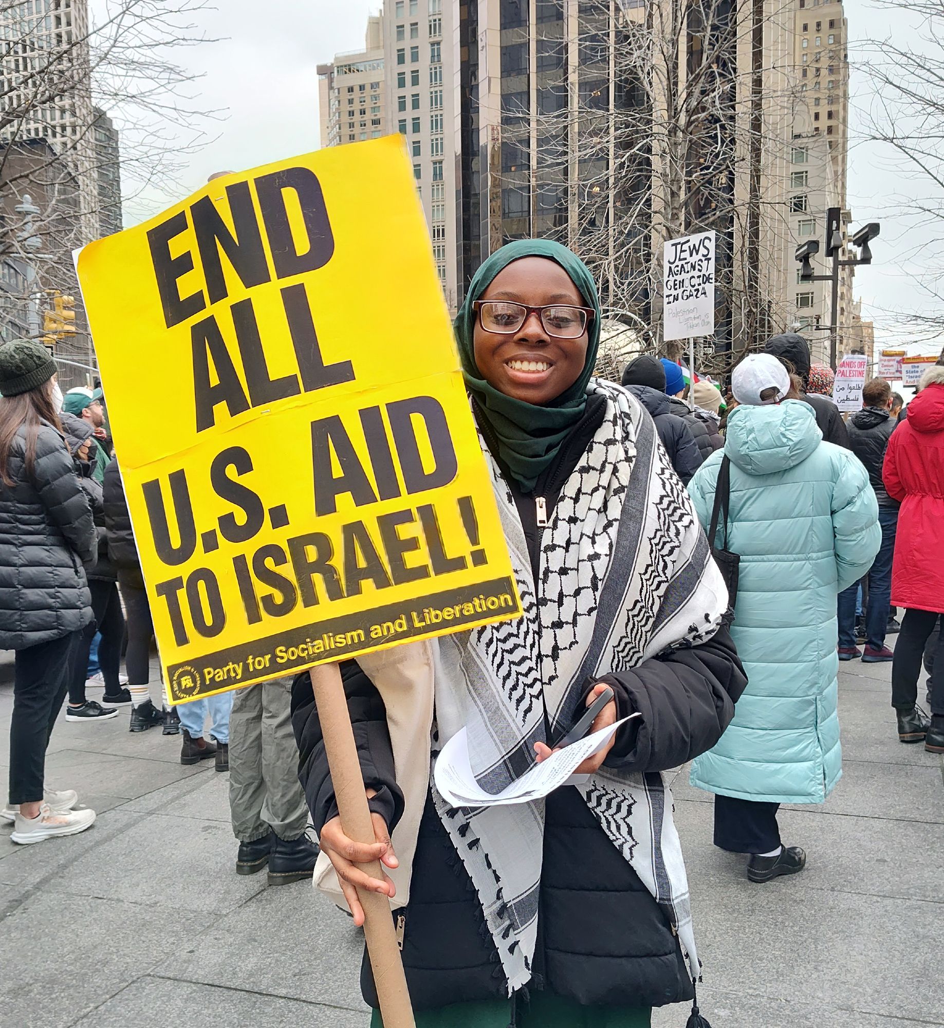 An activist at a protest is wearing a hijab and a keffiyeh holds up a sign saying end all USA to Israel. She is smiling