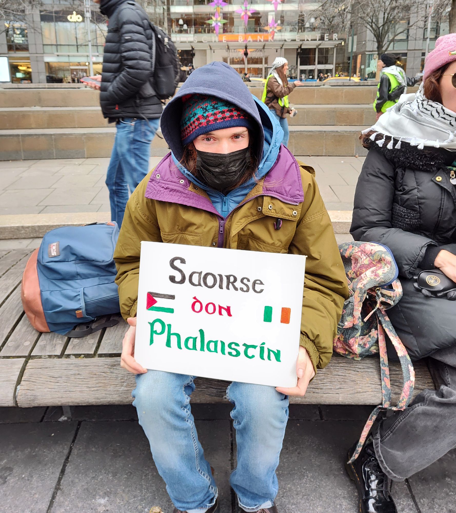 An activist is sitting outside they are bundled up with many layers and their sign is written in Irish so I don't know exactly what they're saying but I do think that the word Palestine is involved and I think they are pro Palestine