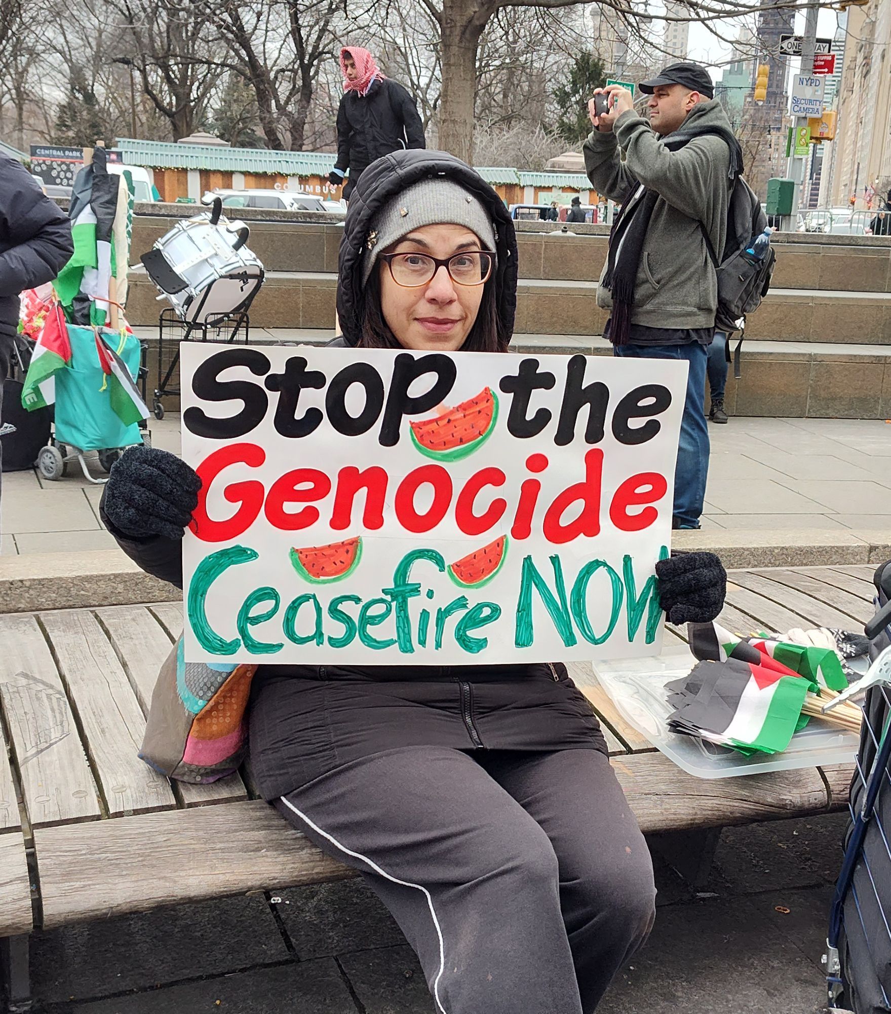 An activist is sitting down before the rally starts and their white sign reads stop the genocide ceasefire now. It is written in marker in black red and green, and they have drawn watermelon symbols