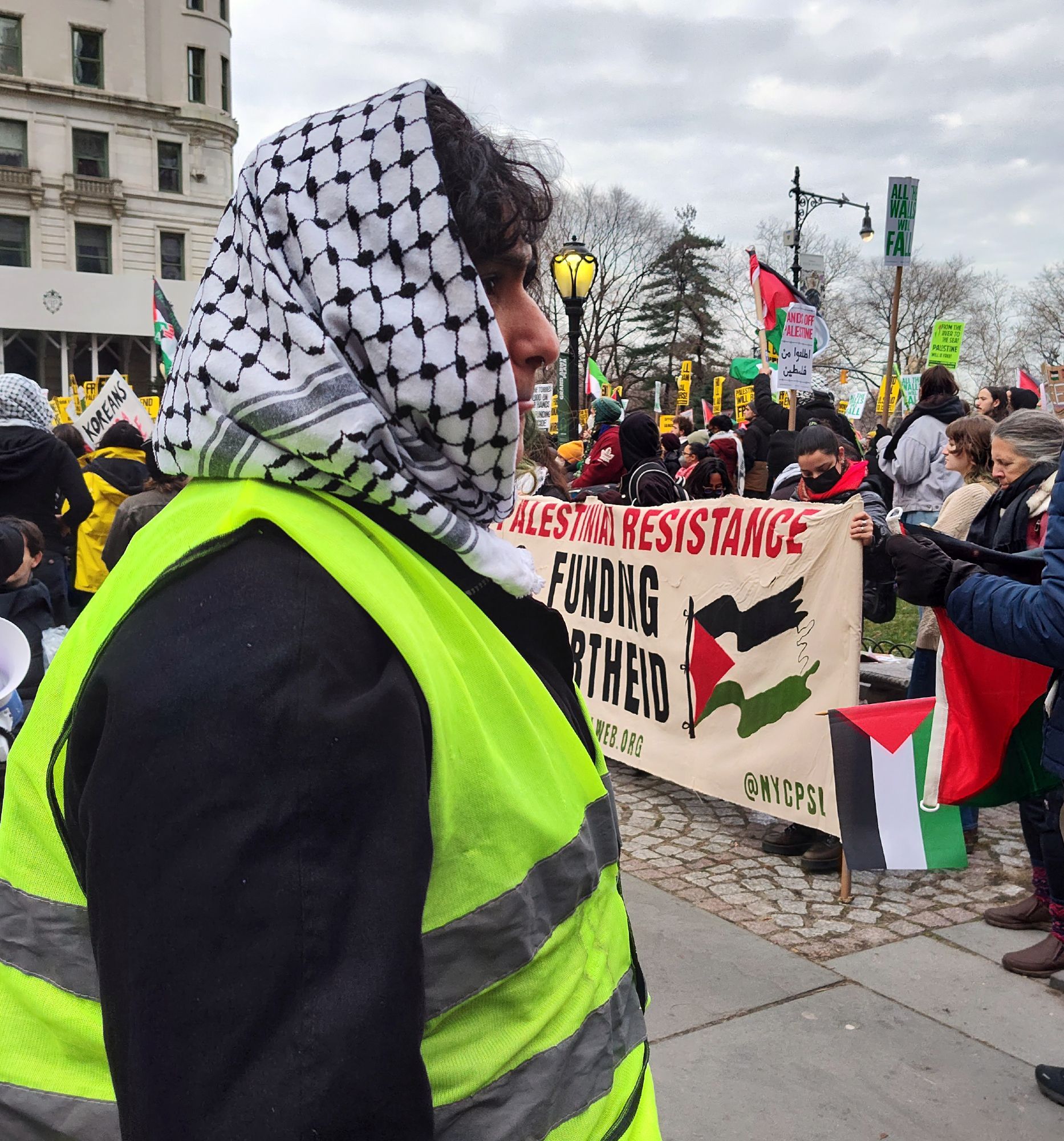 a photo at a NYC protest. the sky is cloudy it's winter. you can see a profile shot of a volunteer. neon yellow green vest. black underneath. they are wearing a keffiyehs. beyond the volunteer (who was very nice) you can see a lot of protesters gathered behind a larger banner as people were getting ready to eventually march.