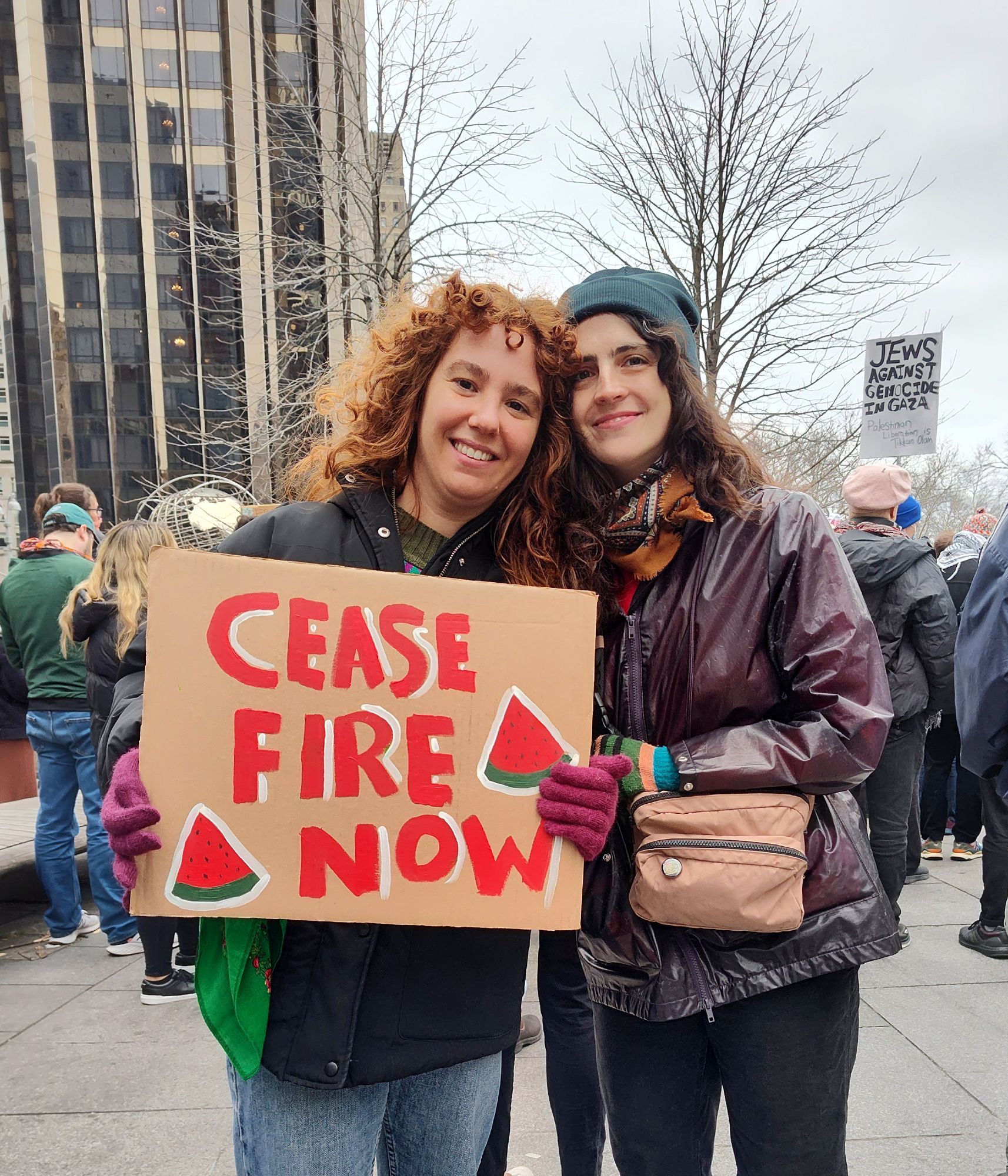 Two activists are outside of a rally New York City. Their heads are close together as one of them holds up a cardboard sign that reads ceasefire now with watermelon symbols on them