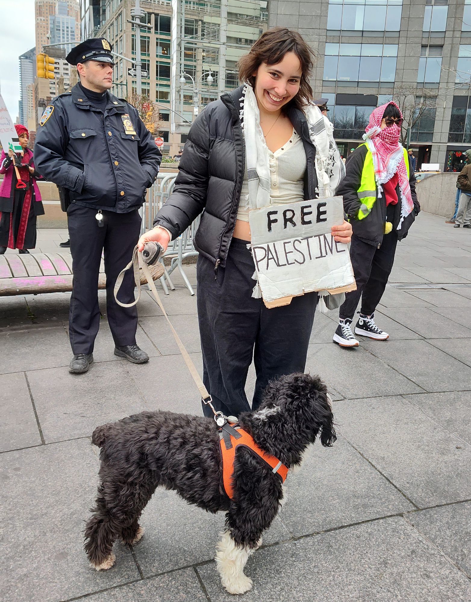 An activist and their dog is at a protest in New York City outside in Columbus circle. She holds up with signs saying free Palestine. There's a cop behind her