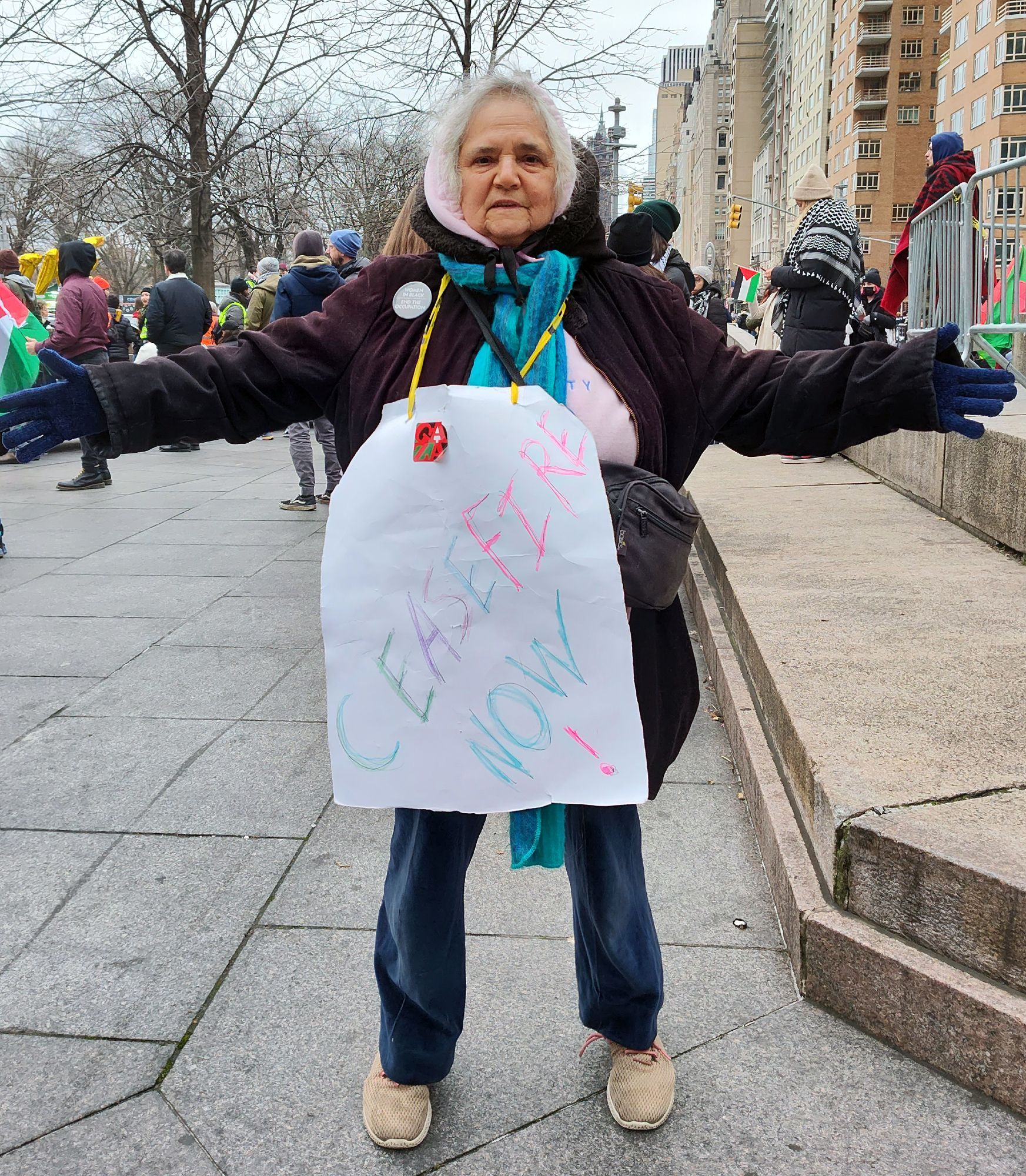 An activist is standing outside of a protest in Columbus circle in New York City. She's bundled up for the weather and her large sign which hangs around her neck reads cease fire now. Her arms are stretched out. She is an older woman and told me that she is a Jewish activist who is anti-Zionist