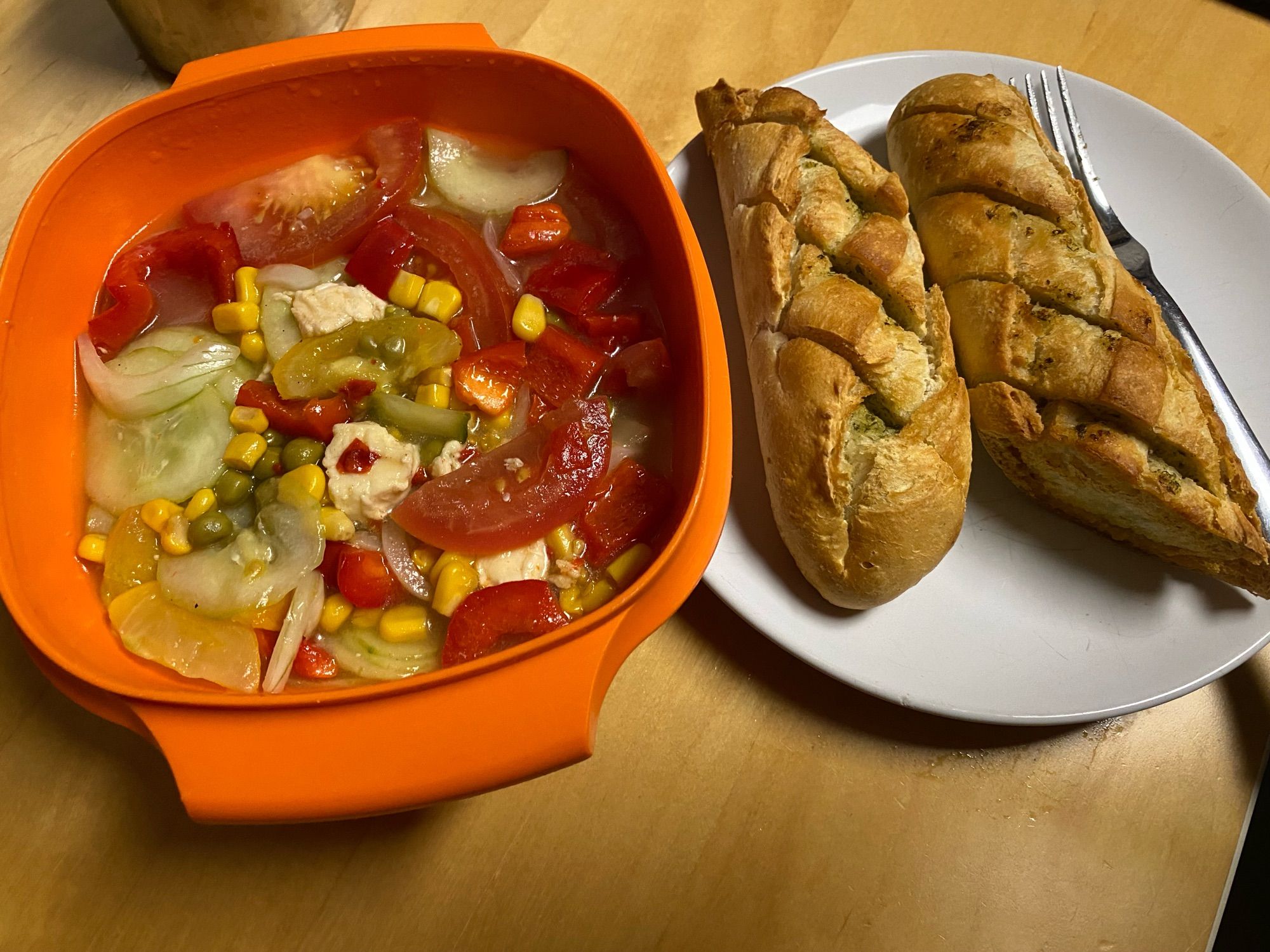 A tupperware storage container with mixed salad - tomatoes, cucumbers, corn, zucchini, bellpepper, mozzarella) next to a plate with an oven baked garlic bread