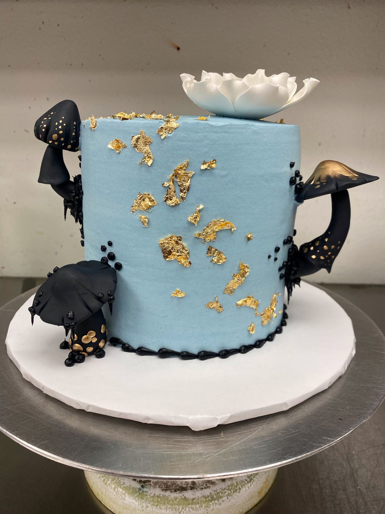 A cake iced in light blue frosting with three black and gold mushroom decorations around it. Gold flakes along the side and a black border on the bottom. A white fondant flower sits on top.