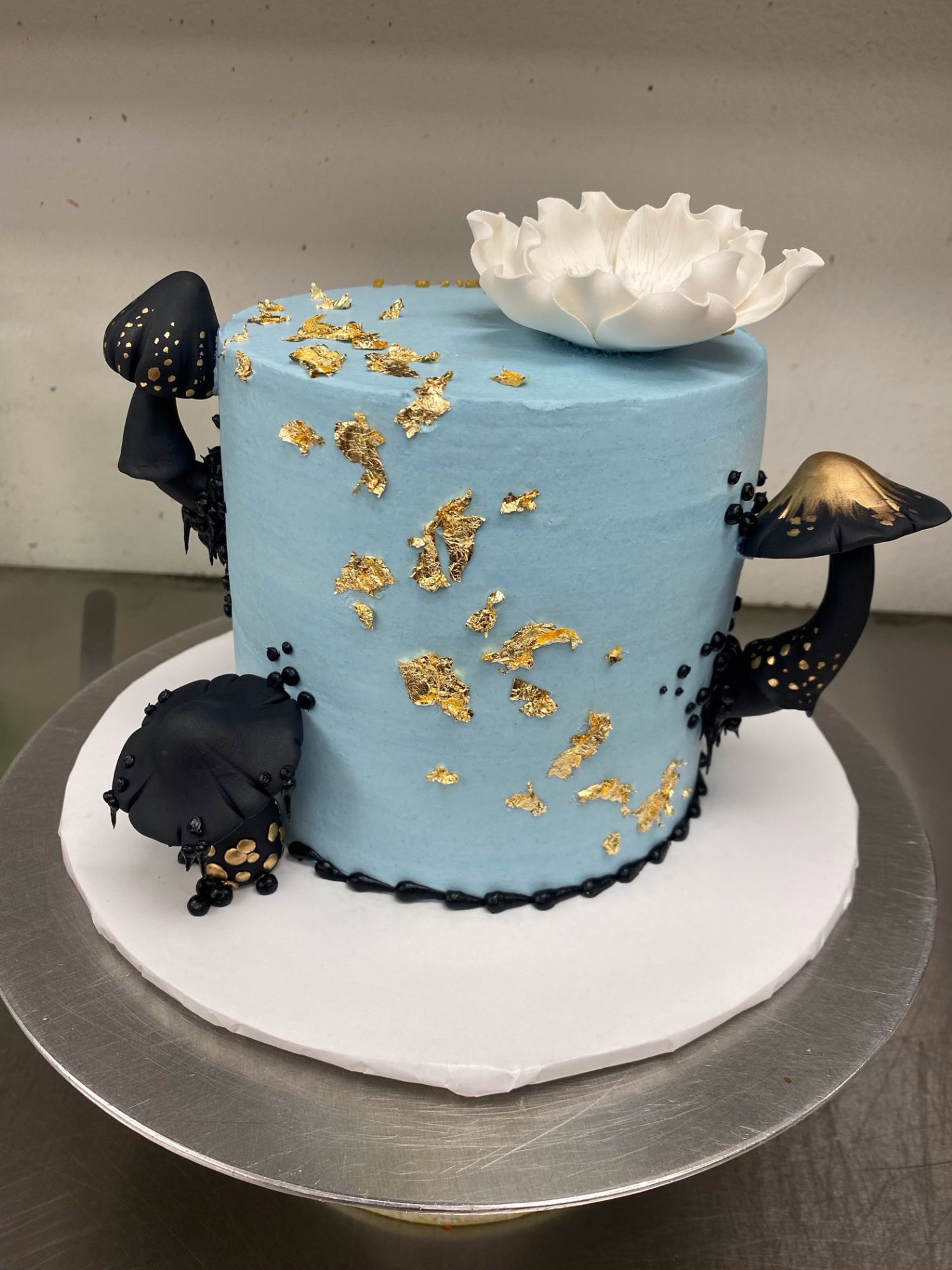 A cake iced in light blue frosting with three black and gold mushroom decorations around it. Gold flakes along the side and a black border on the bottom. A white fondant flower sits on top.