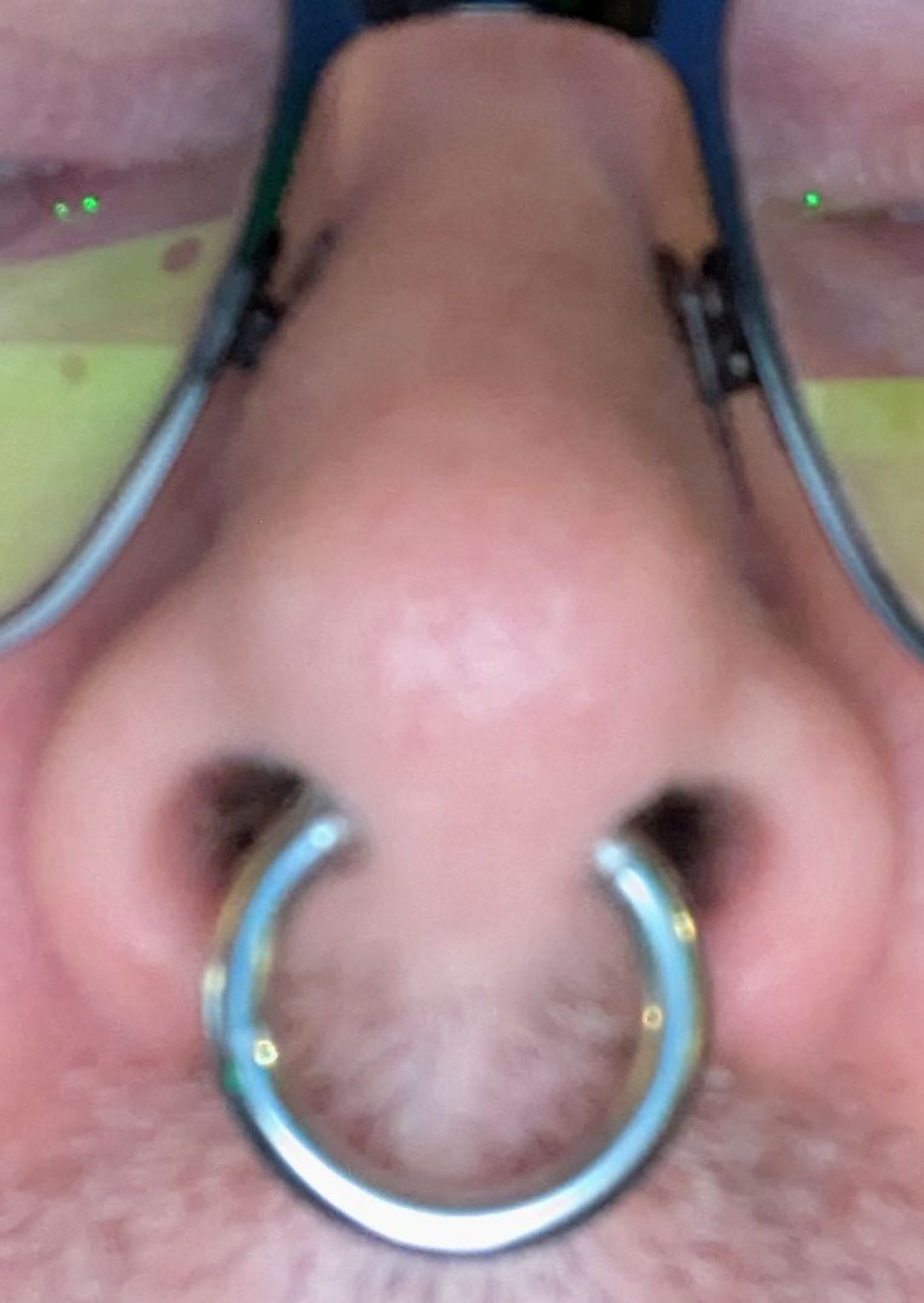 Bildausschnitt einer Nase, in der ein silberner Septum-Piercing-Ring hängt. Die Person trägt eine schwarze Brille und hat ca. 1mm Bart unter der Nase