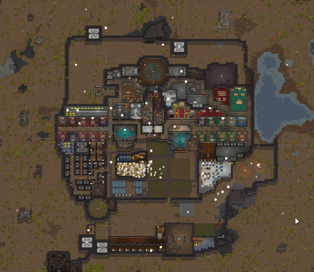 A different Rimworld colony in a less-green area. Prisoner barracks are to the south by a fighting chokepoint, and well-fortified black walls surround the area. There's a church at a central point in the building. There are also an alarming amount of special technology pods. Praise the robot cult!