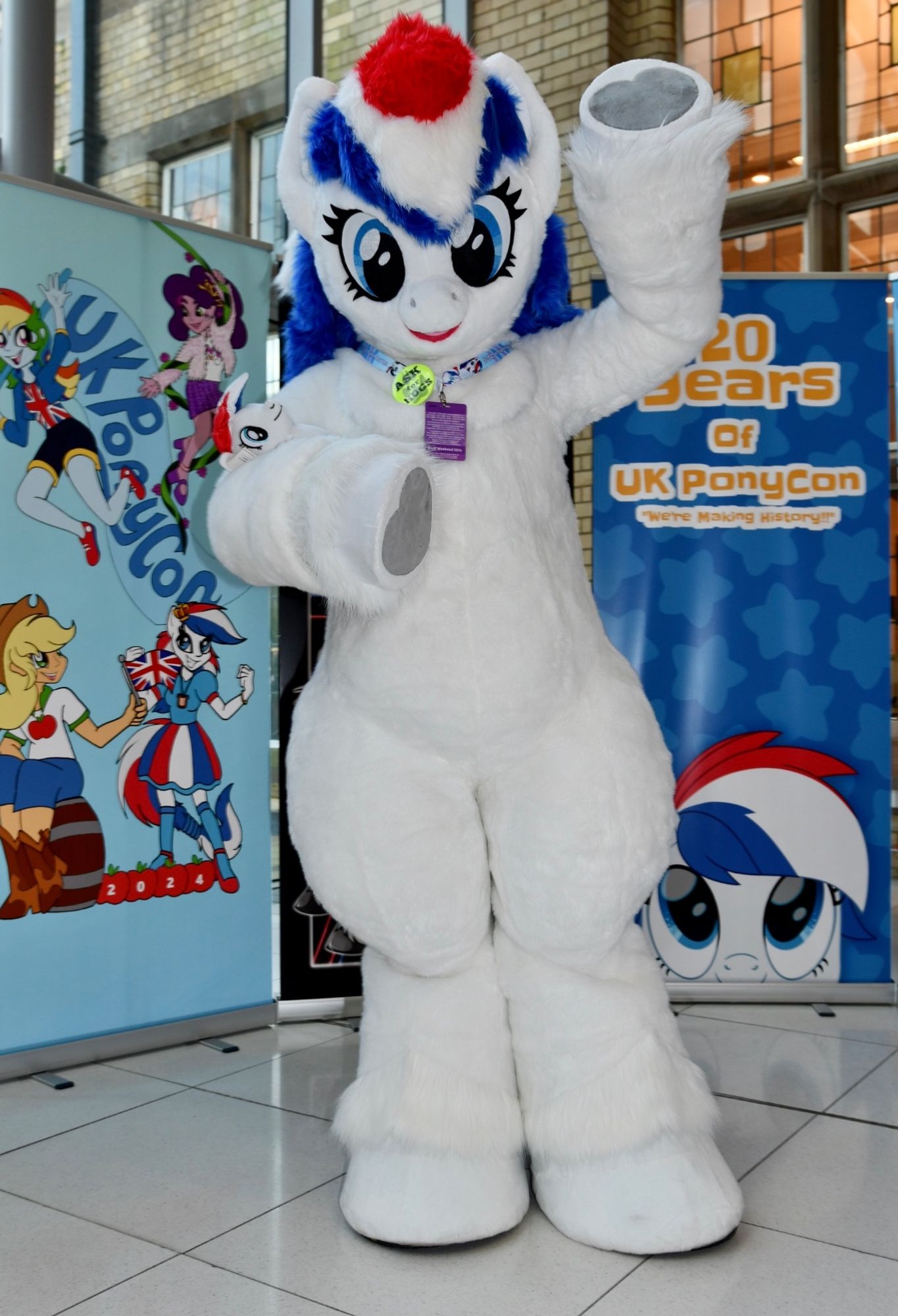 A fursuit of a white pony with red, white, & blue stripes in her mane. She is waving at the camera with hooves that have a heart shaped pattern. There are banners for the con behind her.