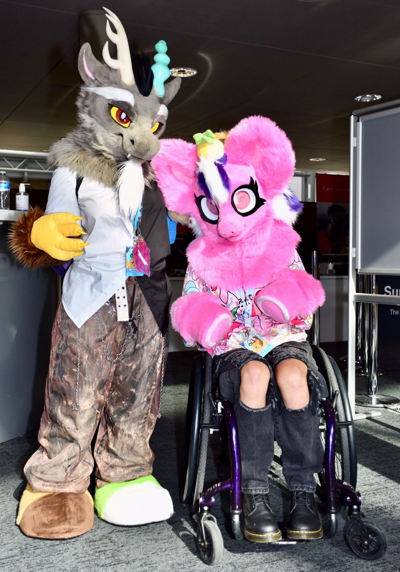 Two fursuiters together. One is based on the character Discord from  ‘My Little Pony: Friendship is Magic’. Discord is a composite creature with brown fur and two different kind of horns. The other is a pink pony with a white & purple mane and large pink eyes.