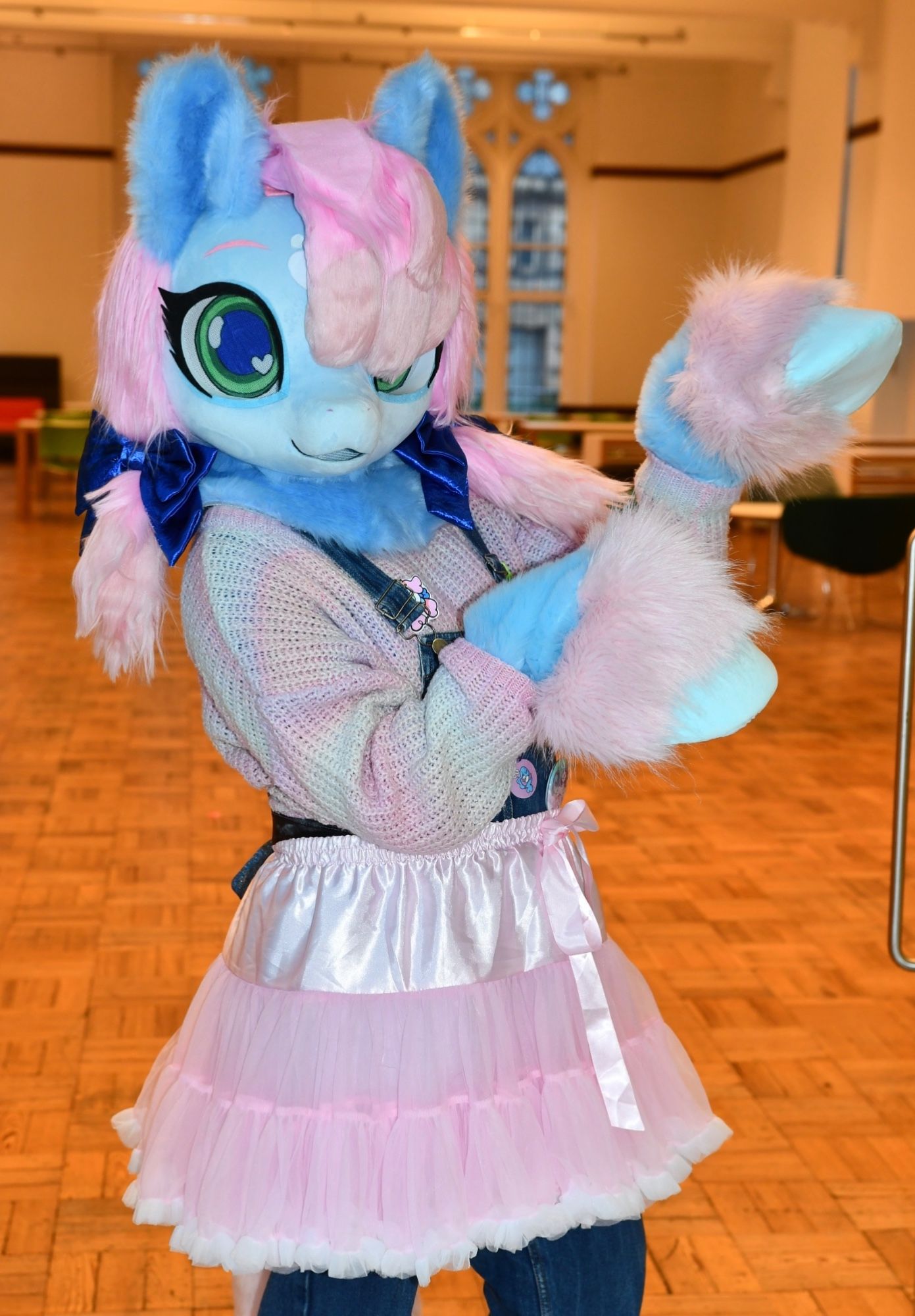 A fursuit of a blue pony with large eyes and a pink mane with dark blue satin bows. She is wearing a pink & light green sweater with a pink tutu.