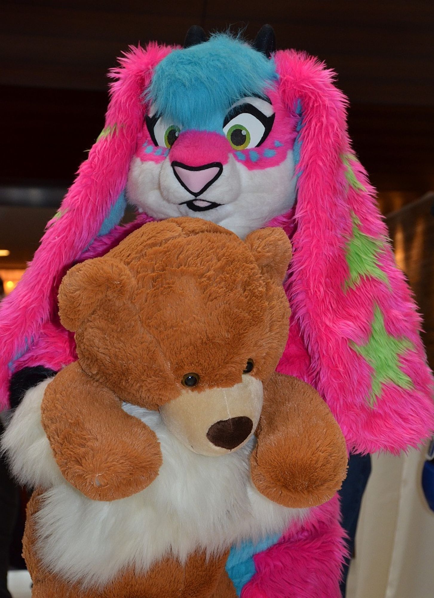 A pink & blue bunny/goat fursuit. She has incredibly long ears with a green star pattern on them. She is holding a large brown teddy bear.