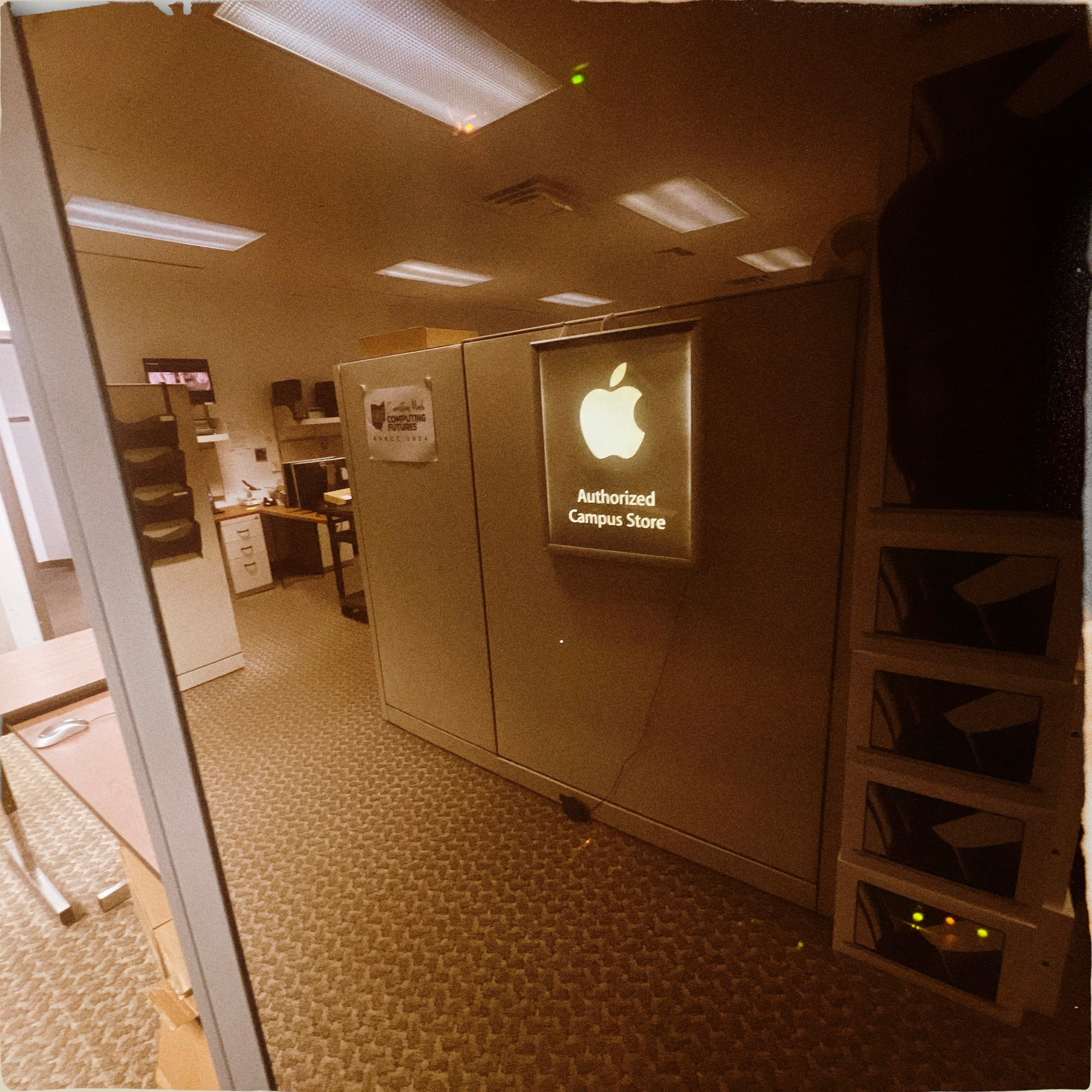 A cubical wall with a lighted sign hanging on it with a white Apple Computer logo and the words Authorized Computer Store below it. 